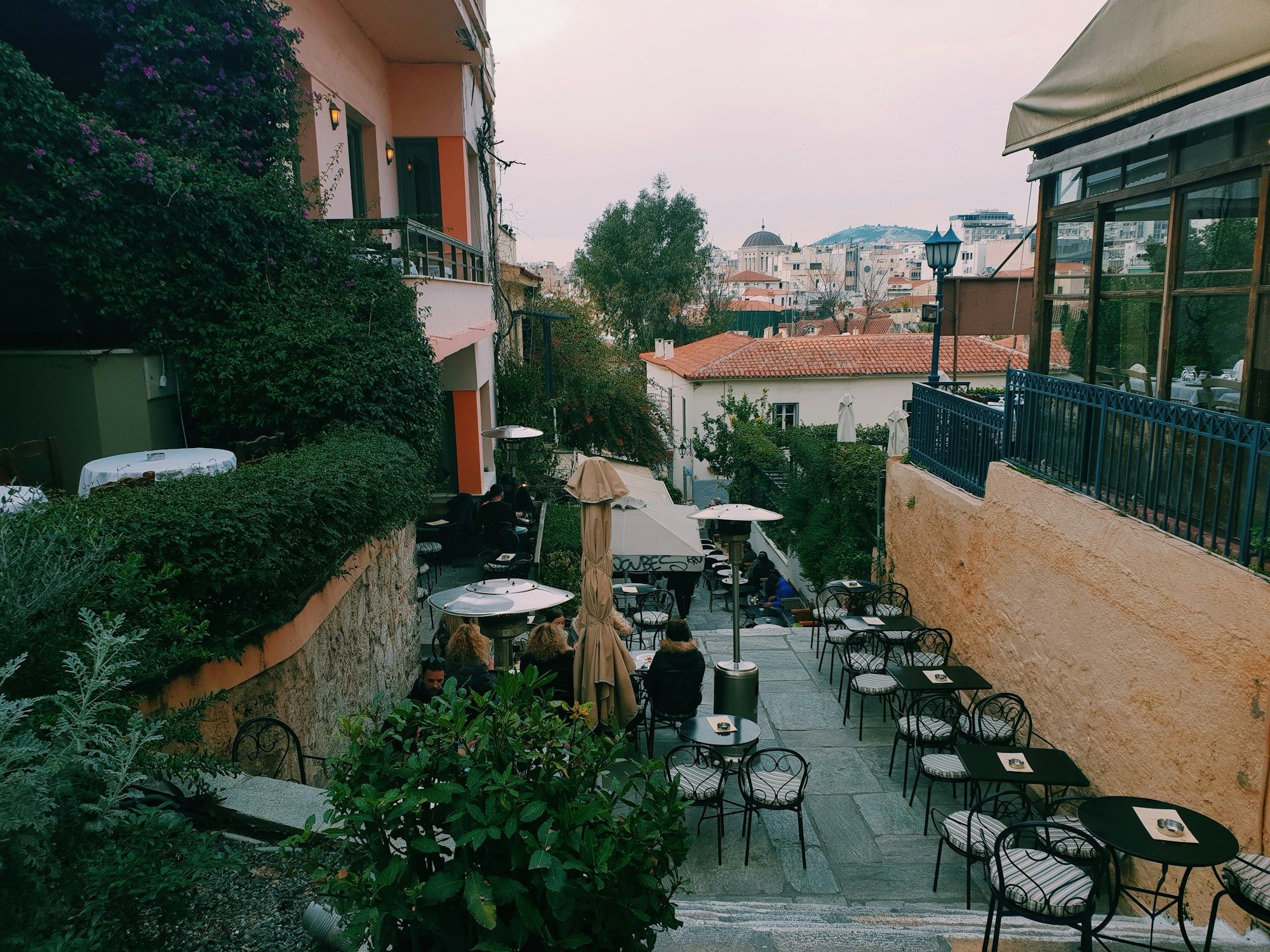 Café tables along a stepped lane in Plaka, Athens, with a few people sitting under umbrellas.