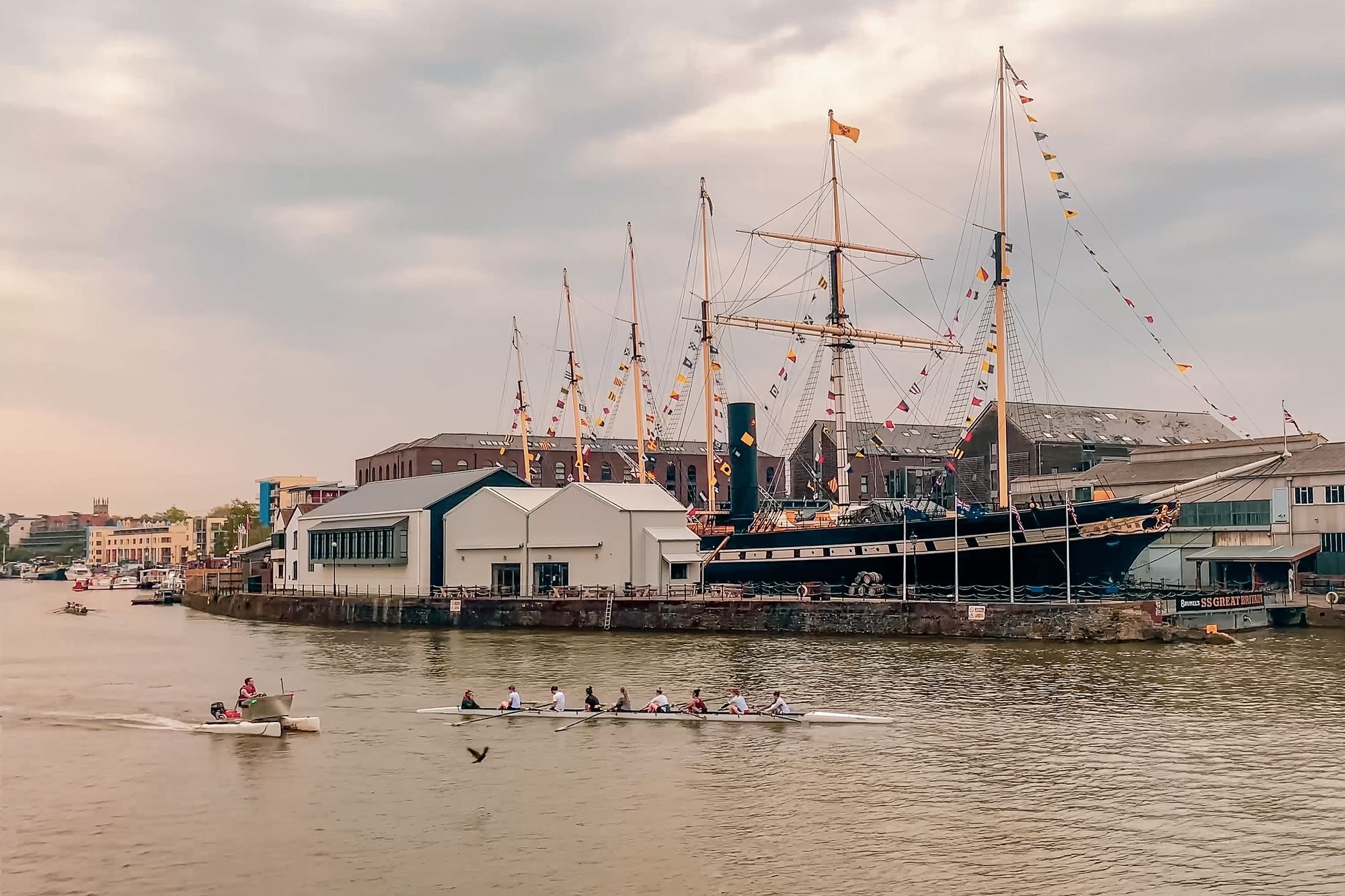 The SS Great Britain moored at Bristol's harbourside, with a rowing crew on the water in front.