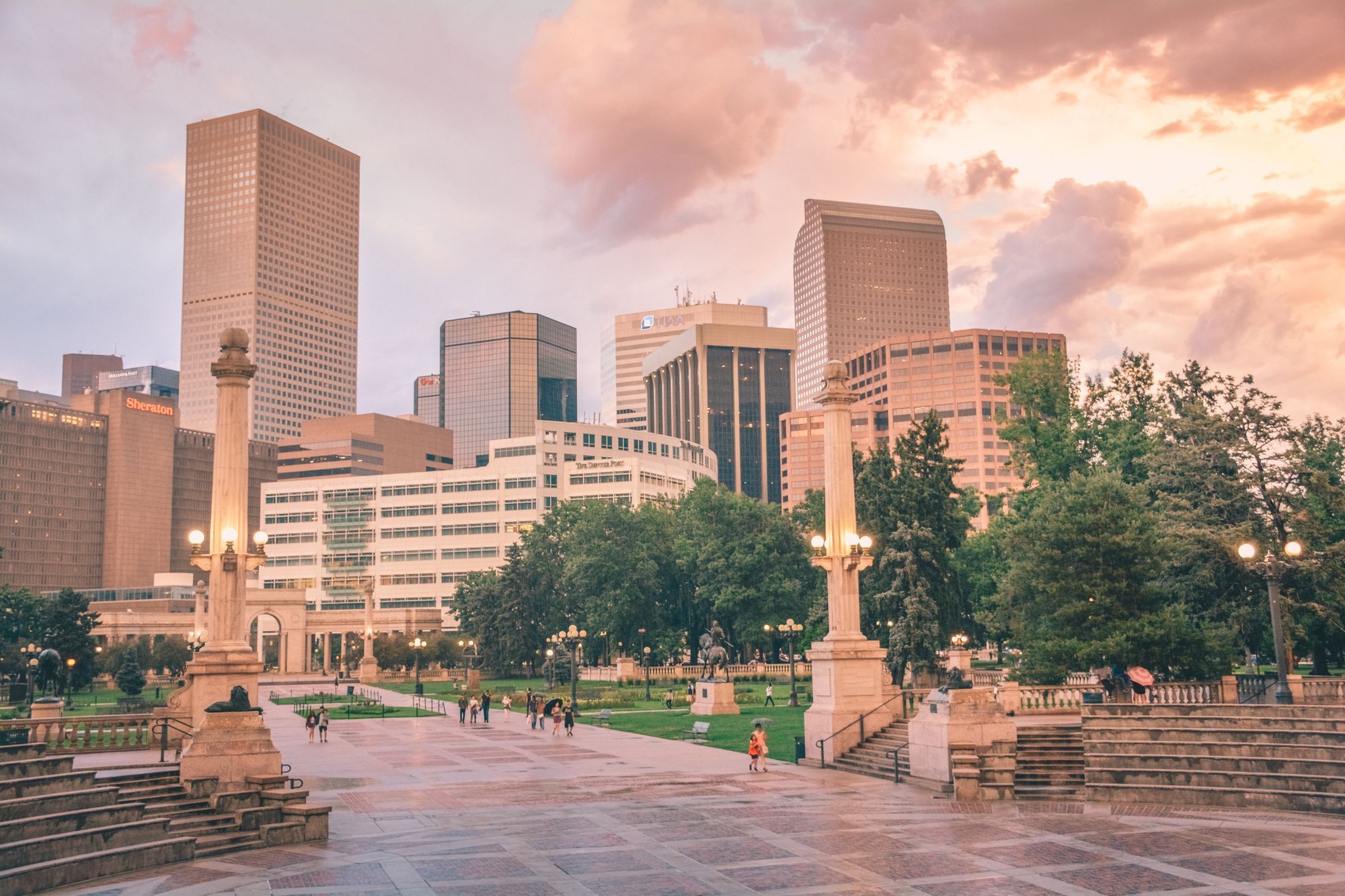 Downtown Denver towers rising behind Civic Center Park at sunset, with people walking across the paved plaza
