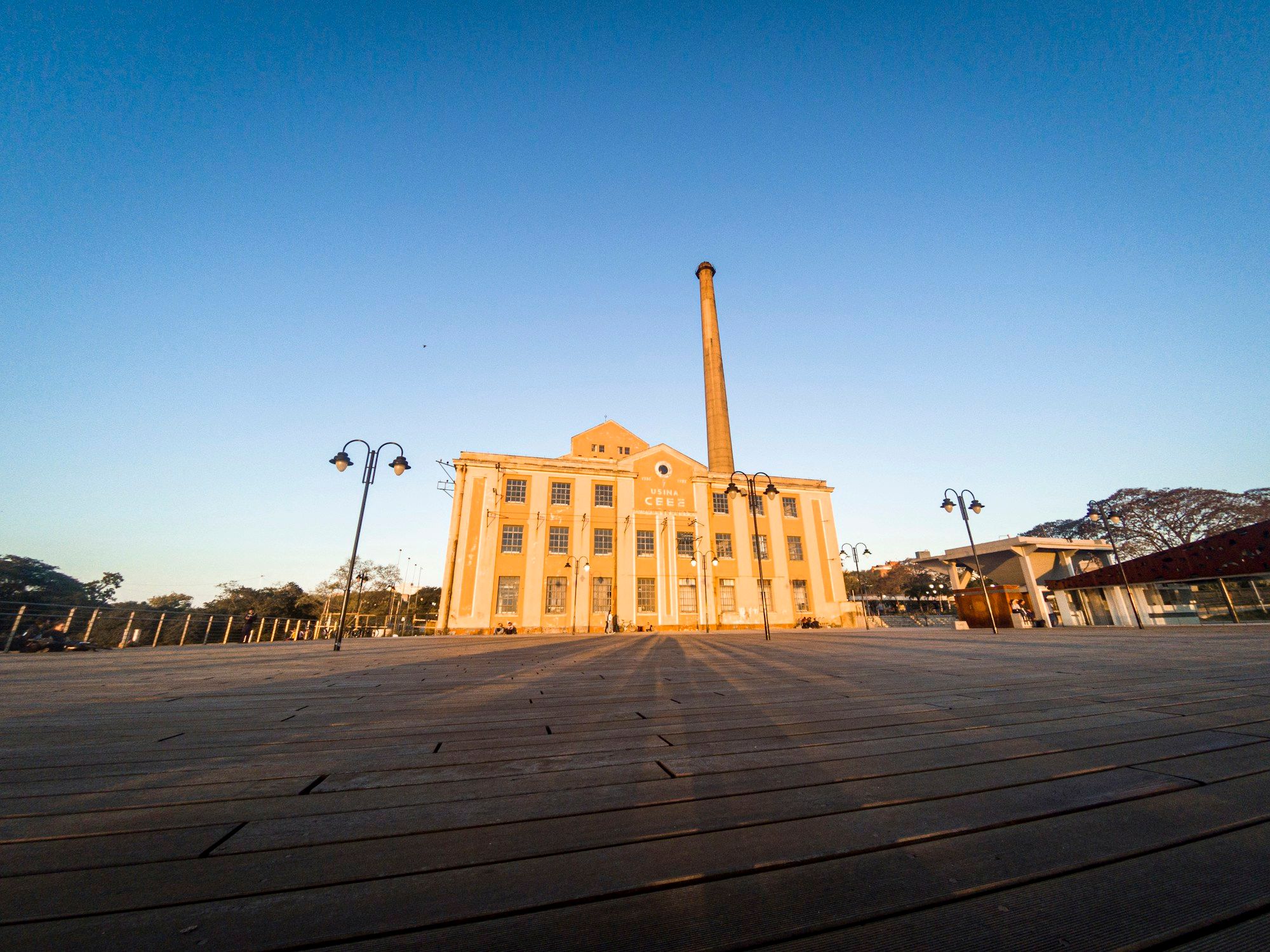 A Usina do Gasômetro e sua chaminé alta sob a luz quente do fim da tarde, vistas de um deque de madeira na orla do Guaíba, em Porto Alegre.