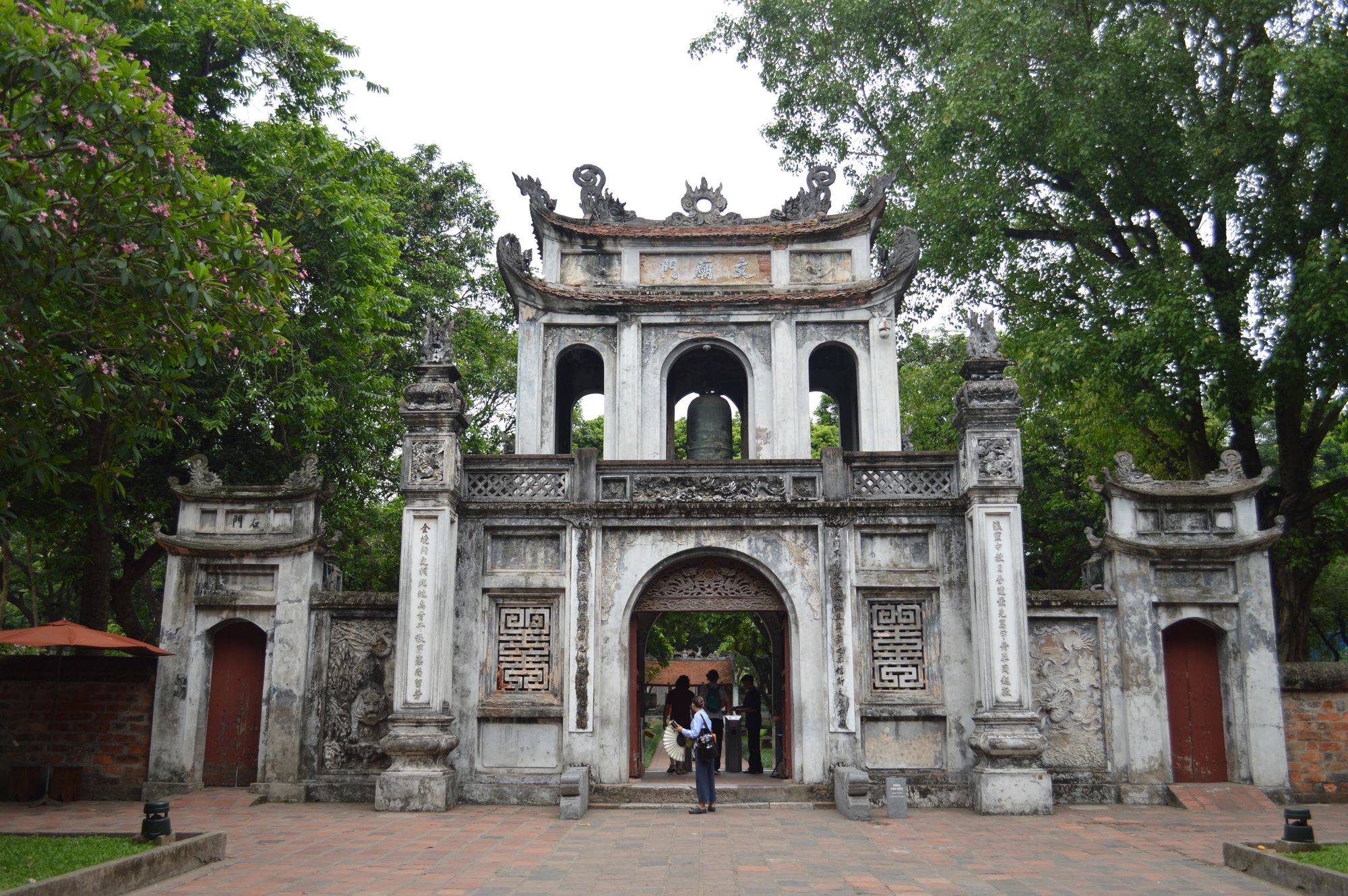 The stone entrance gate of the Temple of Literature in Hanoi, framed by trees, with a paved courtyard in front.