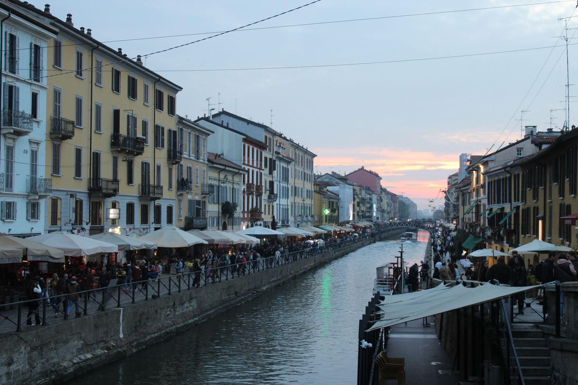 The Naviglio Grande canal in Milan at dusk, with cafe tables and people along the bank and a pink sky above the far end.