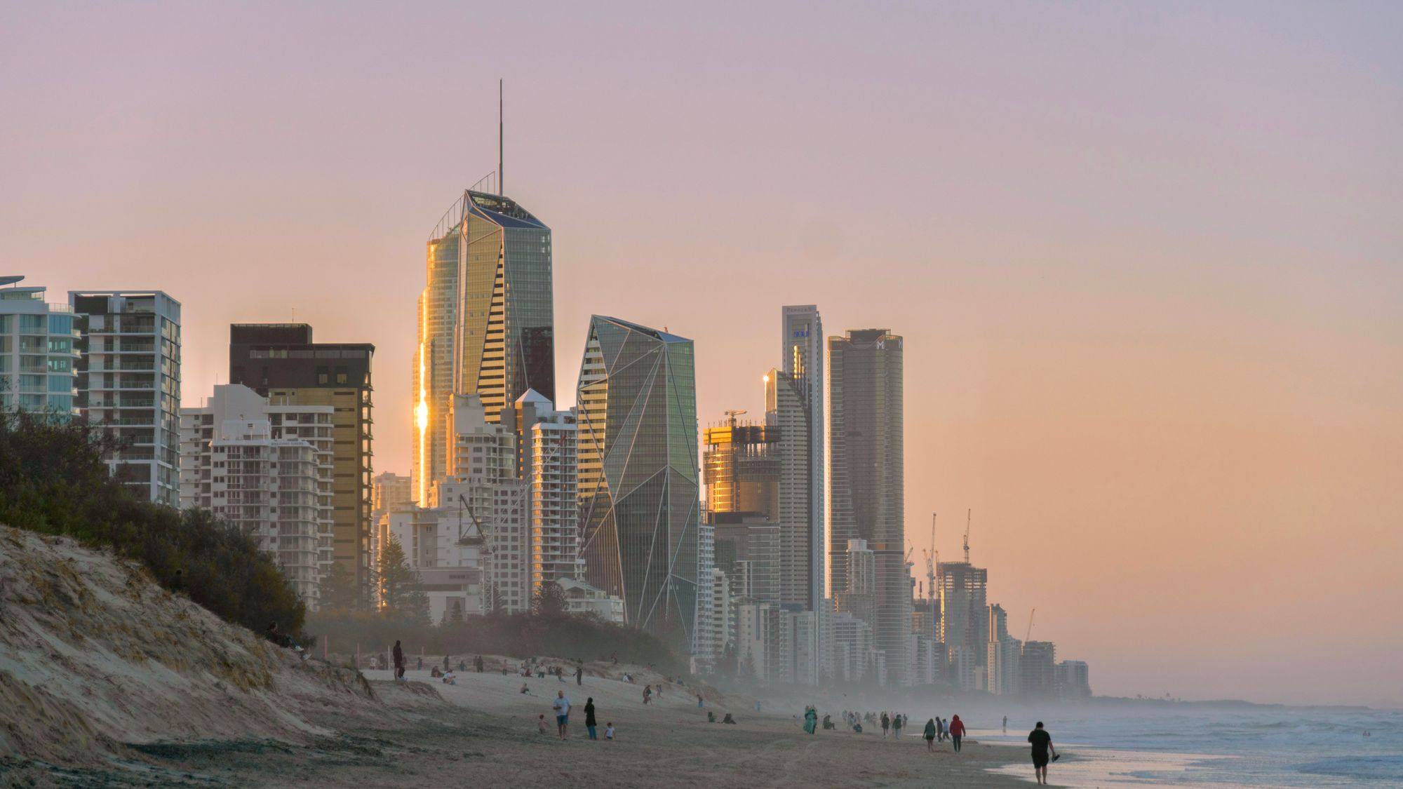 The Surfers Paradise skyline glowing at sunset above the beach on the Gold Coast, with walkers on the sand.