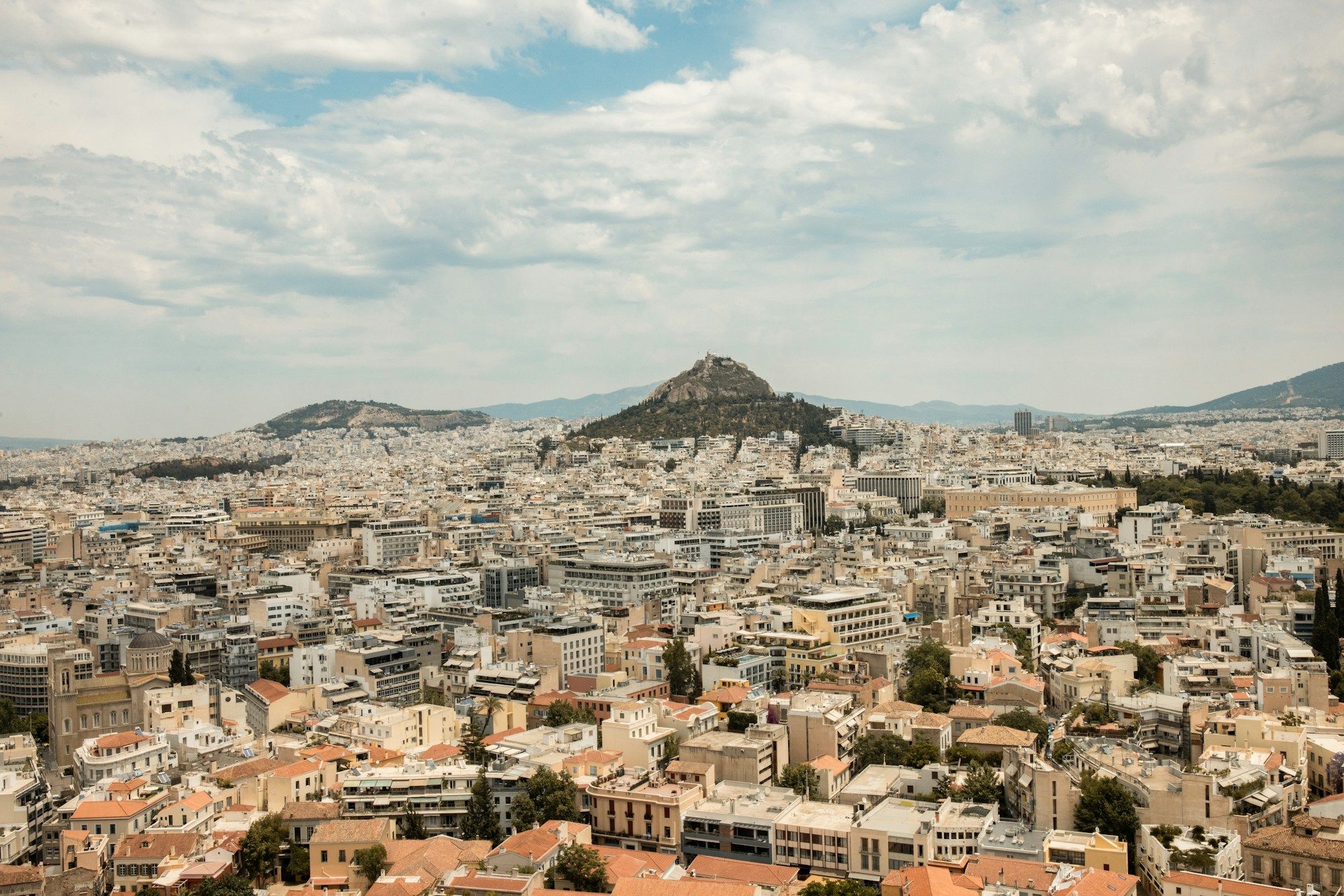 Mount Lycabettus rising above the white buildings of central Athens under a partly cloudy sky.