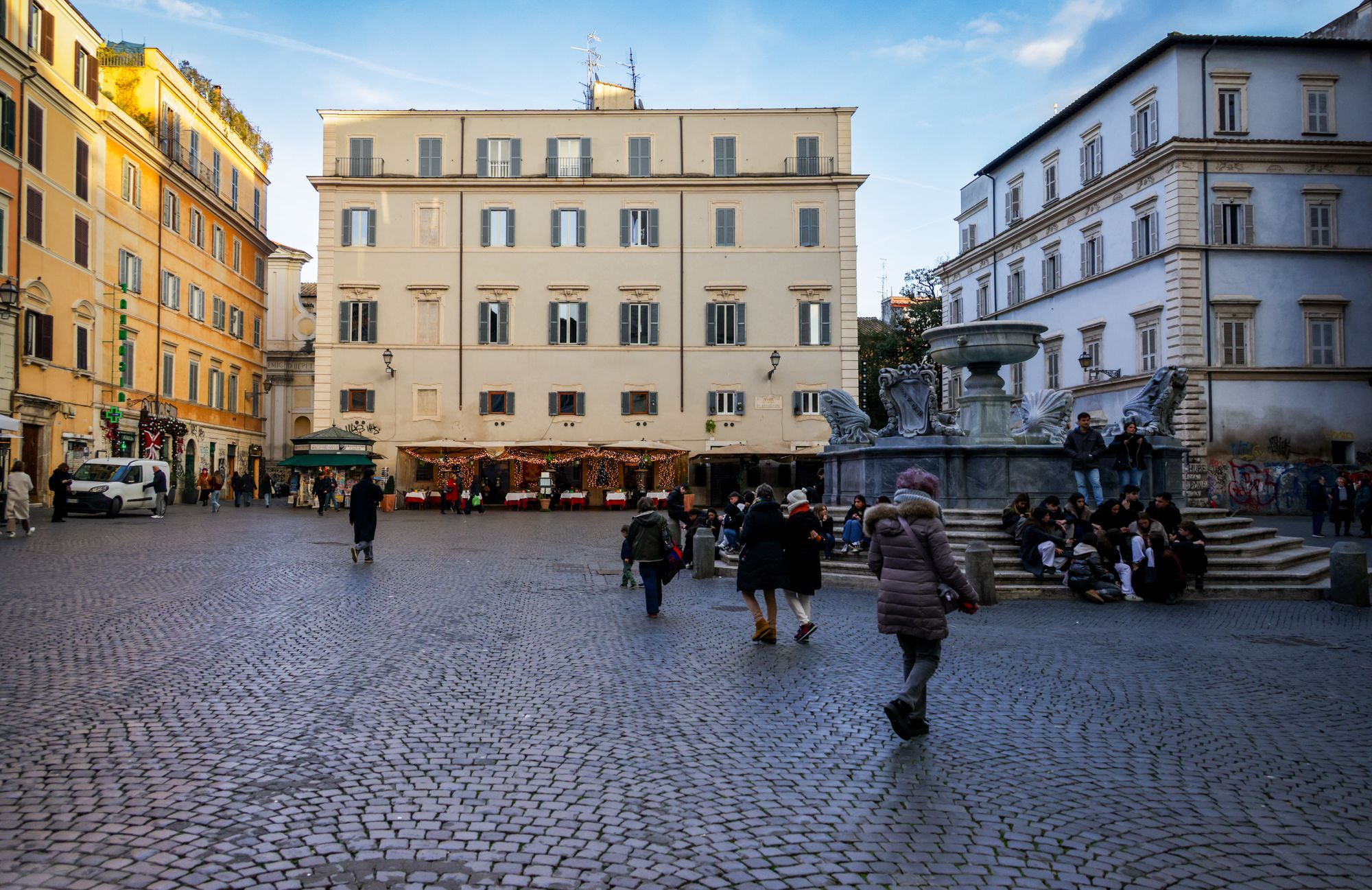 People crossing a cobbled square with a fountain and cafe terraces in Rome's Trastevere.