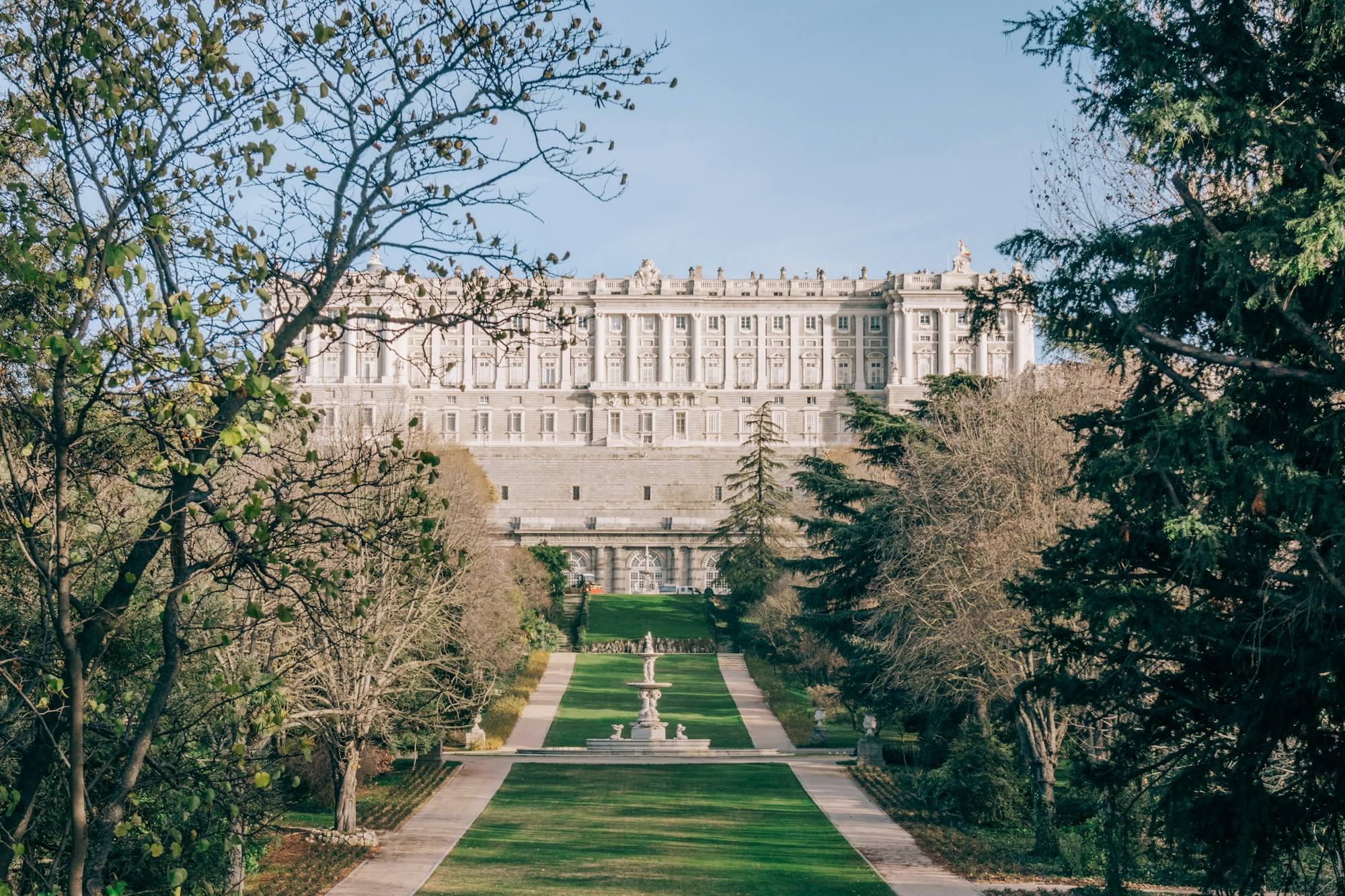 The Royal Palace of Madrid seen from the Campo del Moro gardens, with a lawn avenue and trees in the foreground.