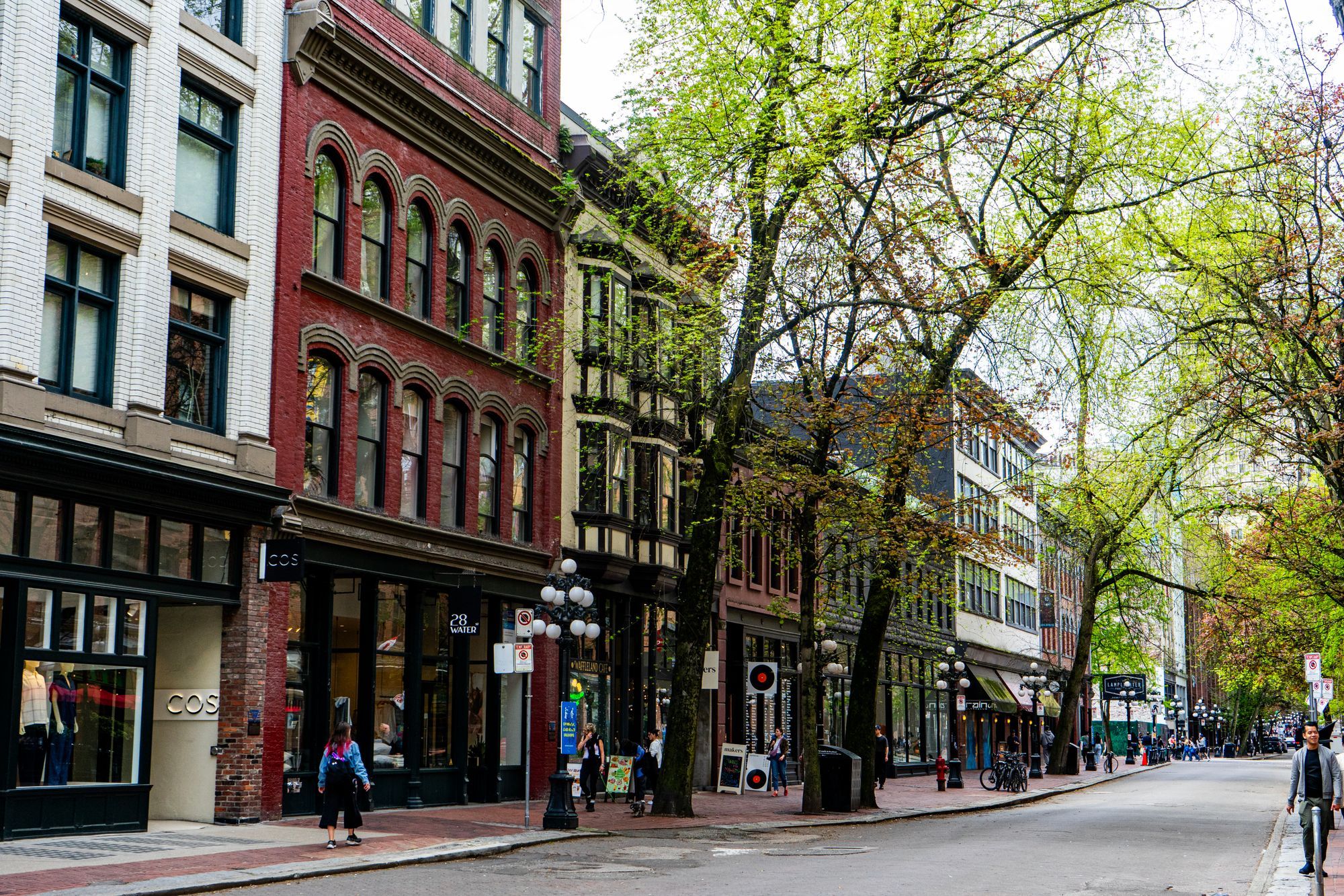 A tree-lined street of brick heritage buildings in Gastown, Vancouver, with a few people walking.