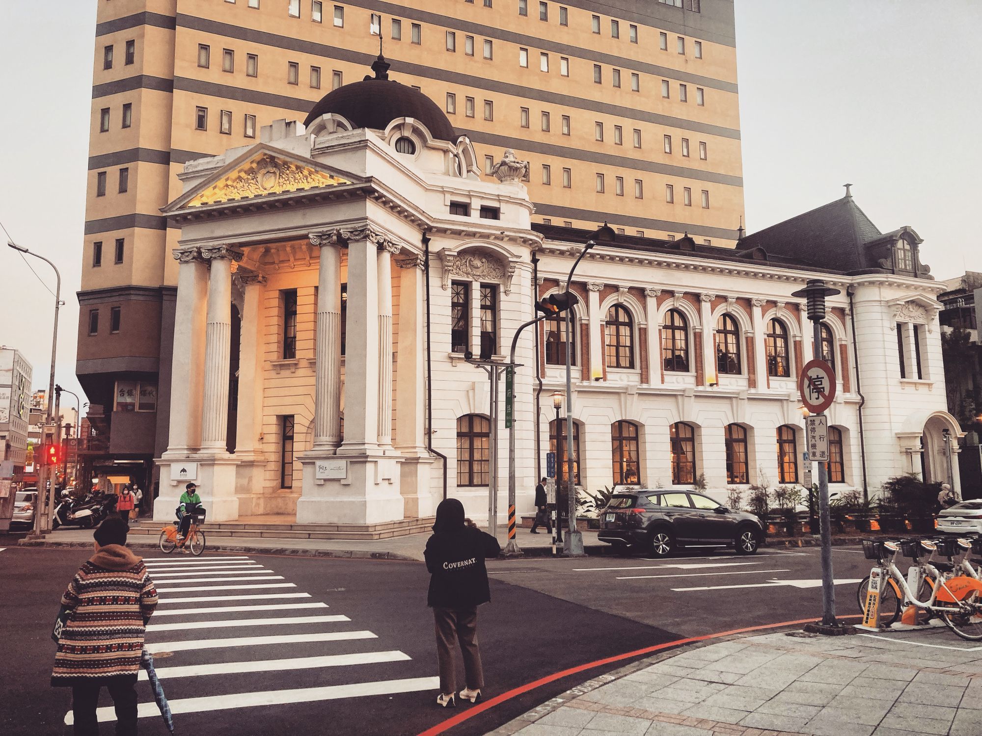 A neoclassical building on a street corner in Taichung at dusk, with people crossing the road in front.