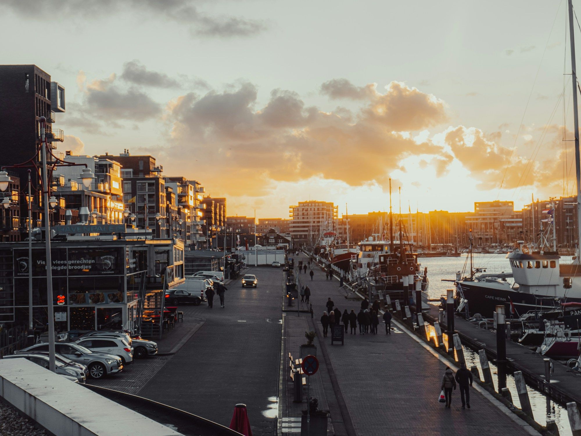 People walking along the quay at Scheveningen harbor in The Hague at sunset, with boats moored on the right.