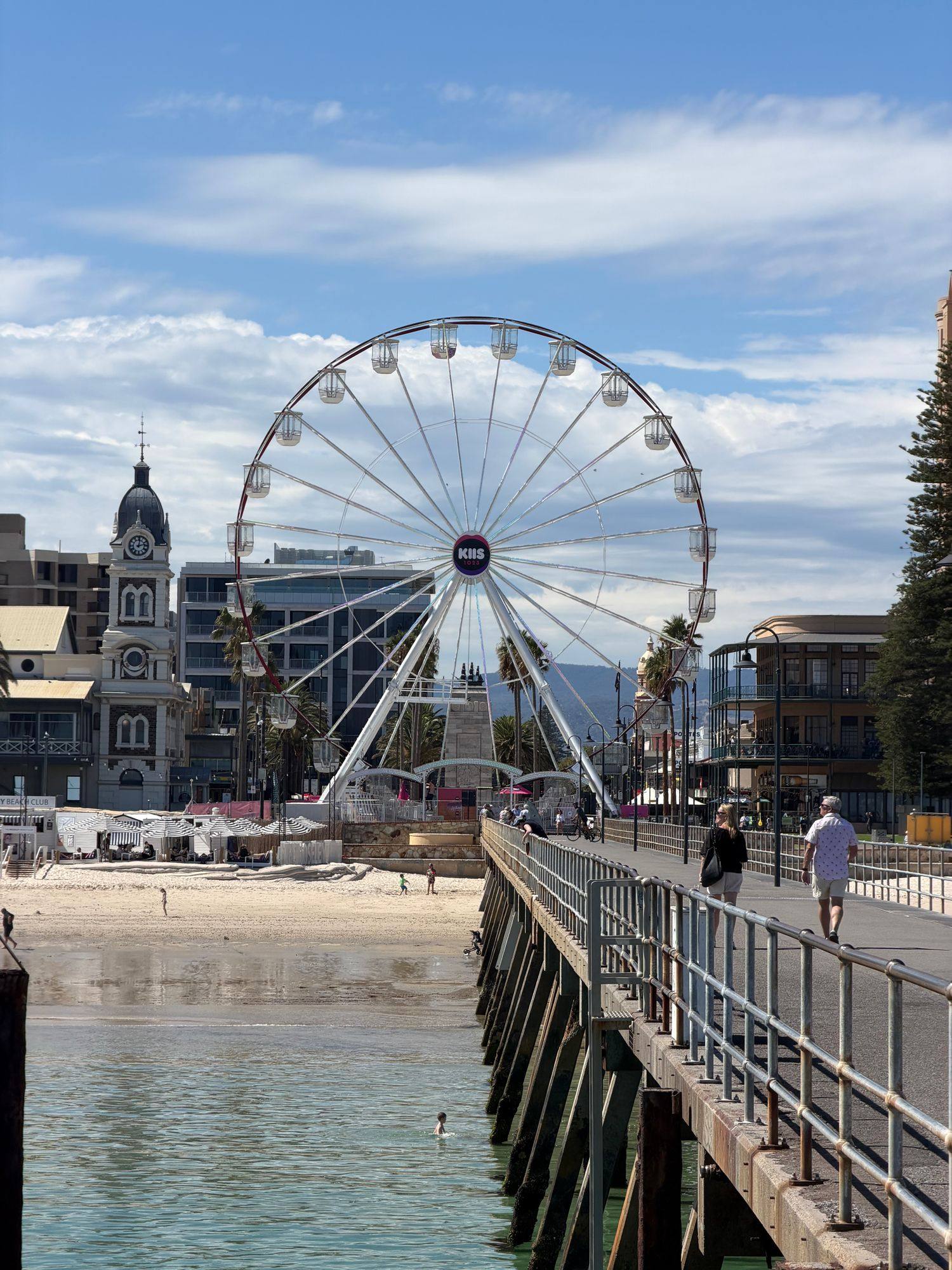The Glenelg Ferris Wheel seen from the pier, with a clock tower and beachfront buildings beside it.