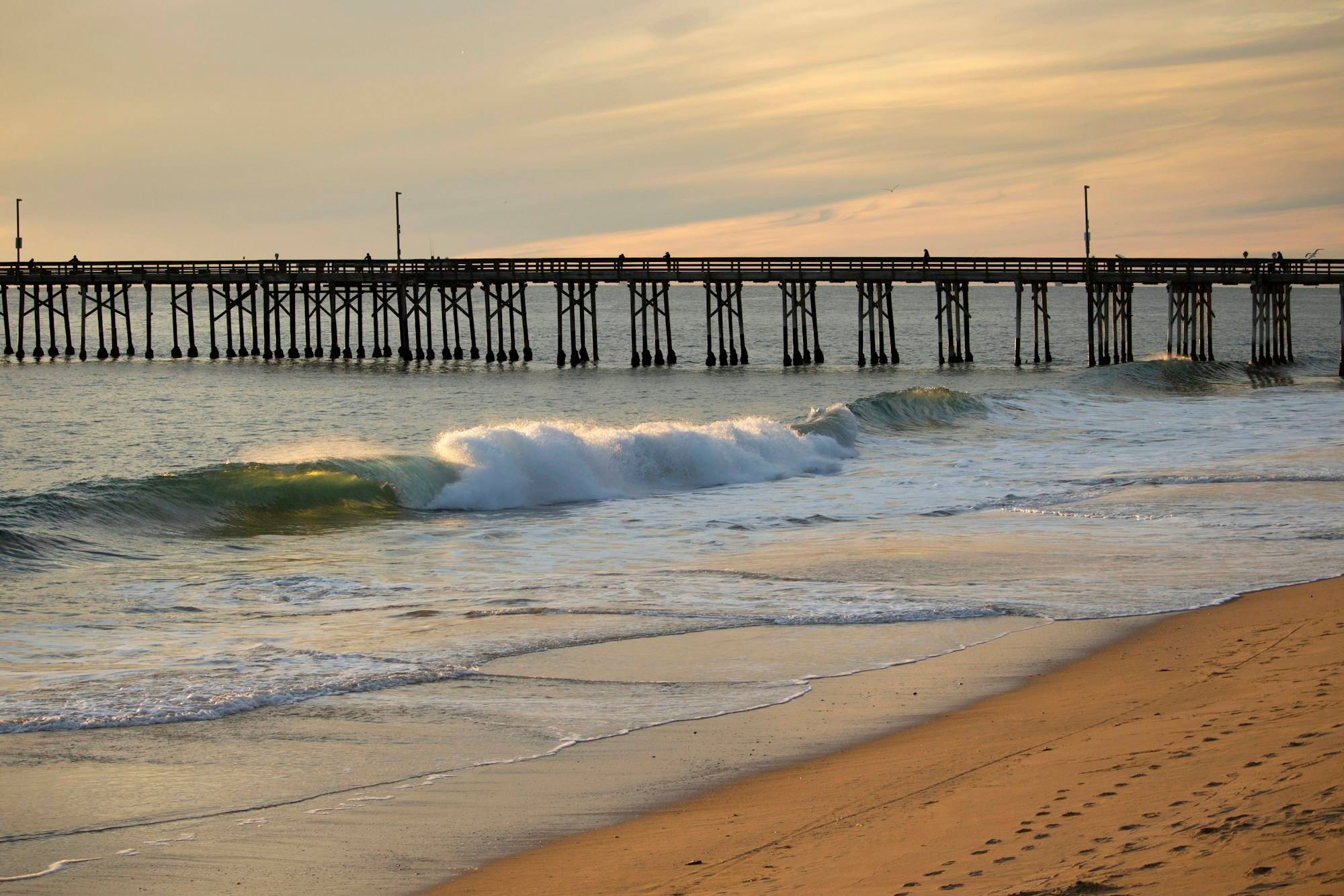 A wooden pier in Newport Beach at sunset, with a wave breaking on the beach in front.