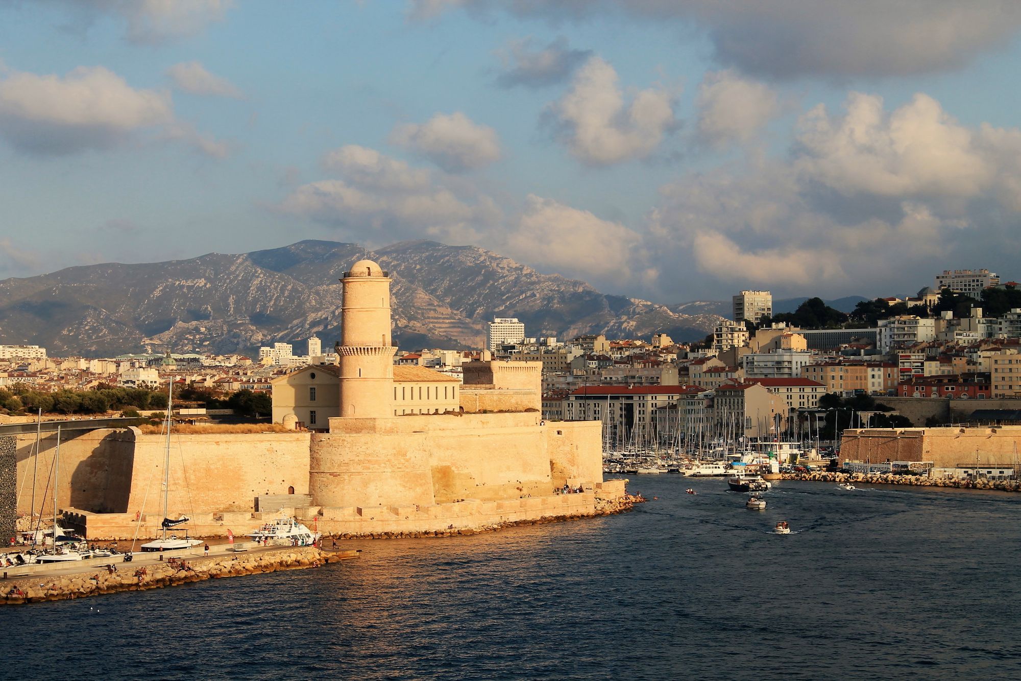 Fort Saint-Jean and its round tower at the entrance of the Vieux-Port in Marseille, in golden evening light with hills behind.