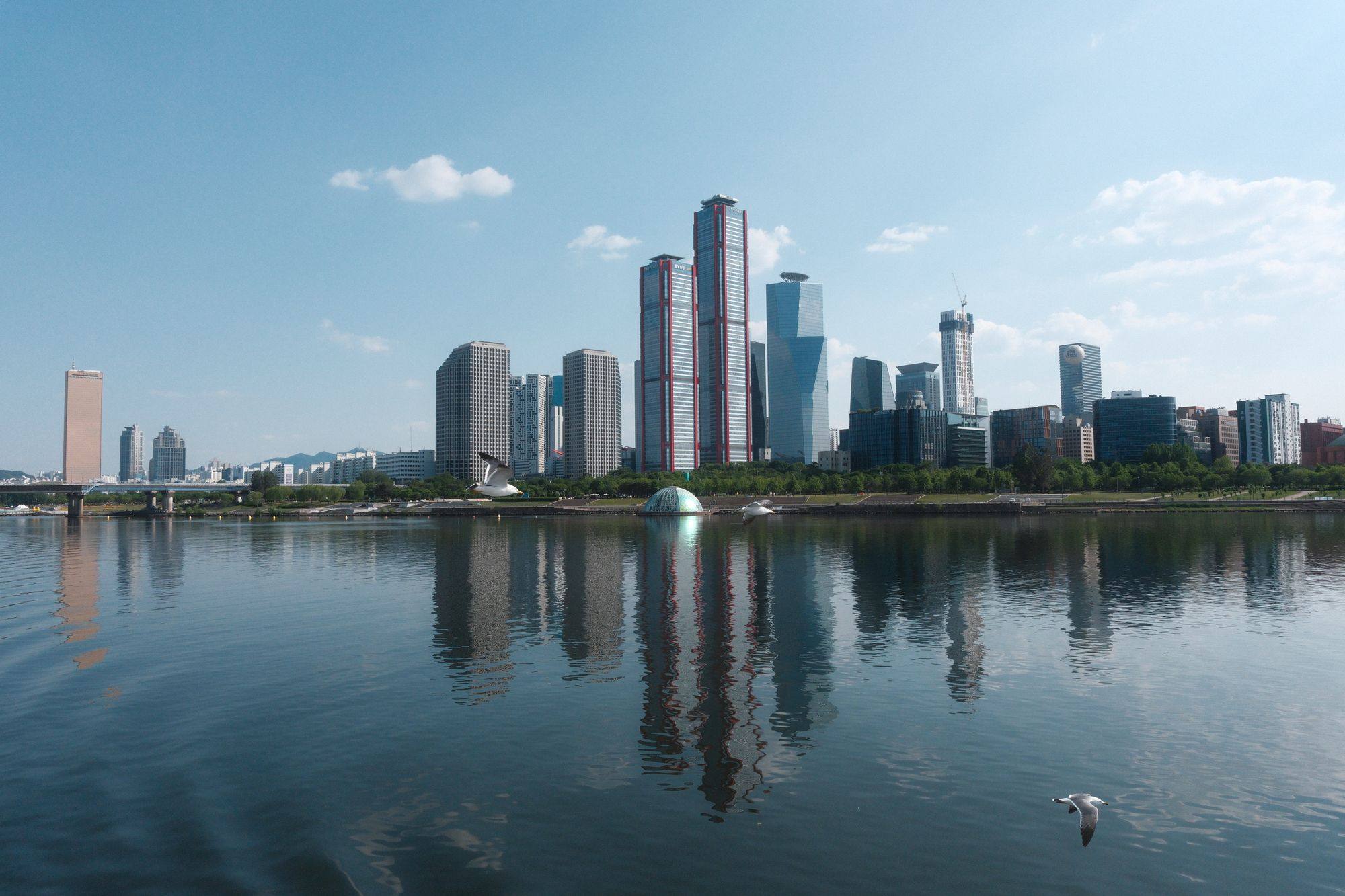 The Seoul skyline of Yeouido across the Han River under a clear sky, with the towers reflected in the water.