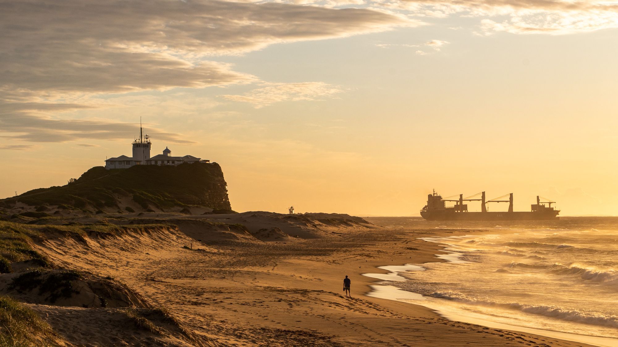 Nobbys Head and its lighthouse above Nobbys Beach in Newcastle at sunset, with a ship on the horizon.