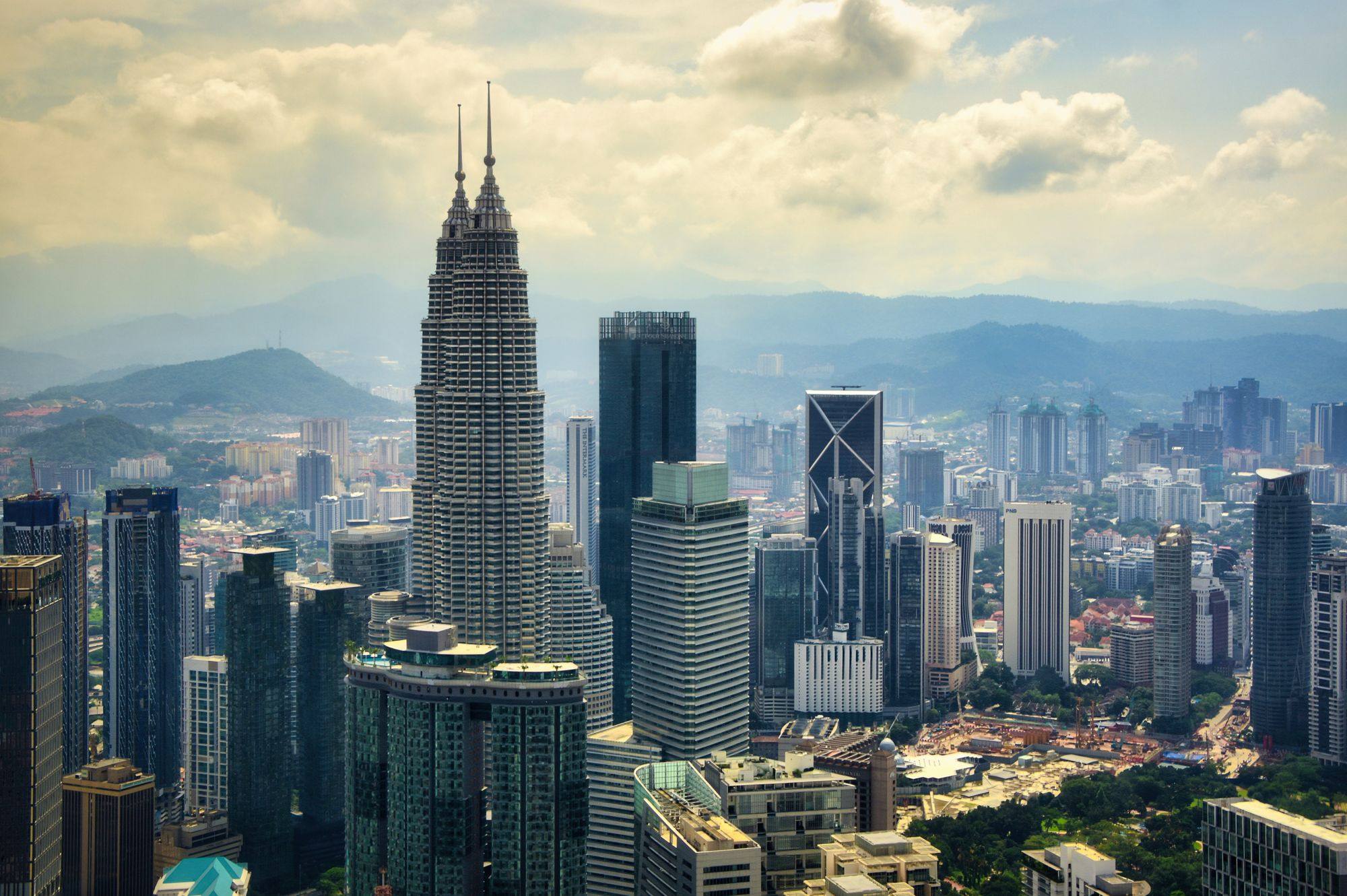 The Petronas Towers rising above the skyline of Kuala Lumpur on a day with soft clouds, with hills behind.