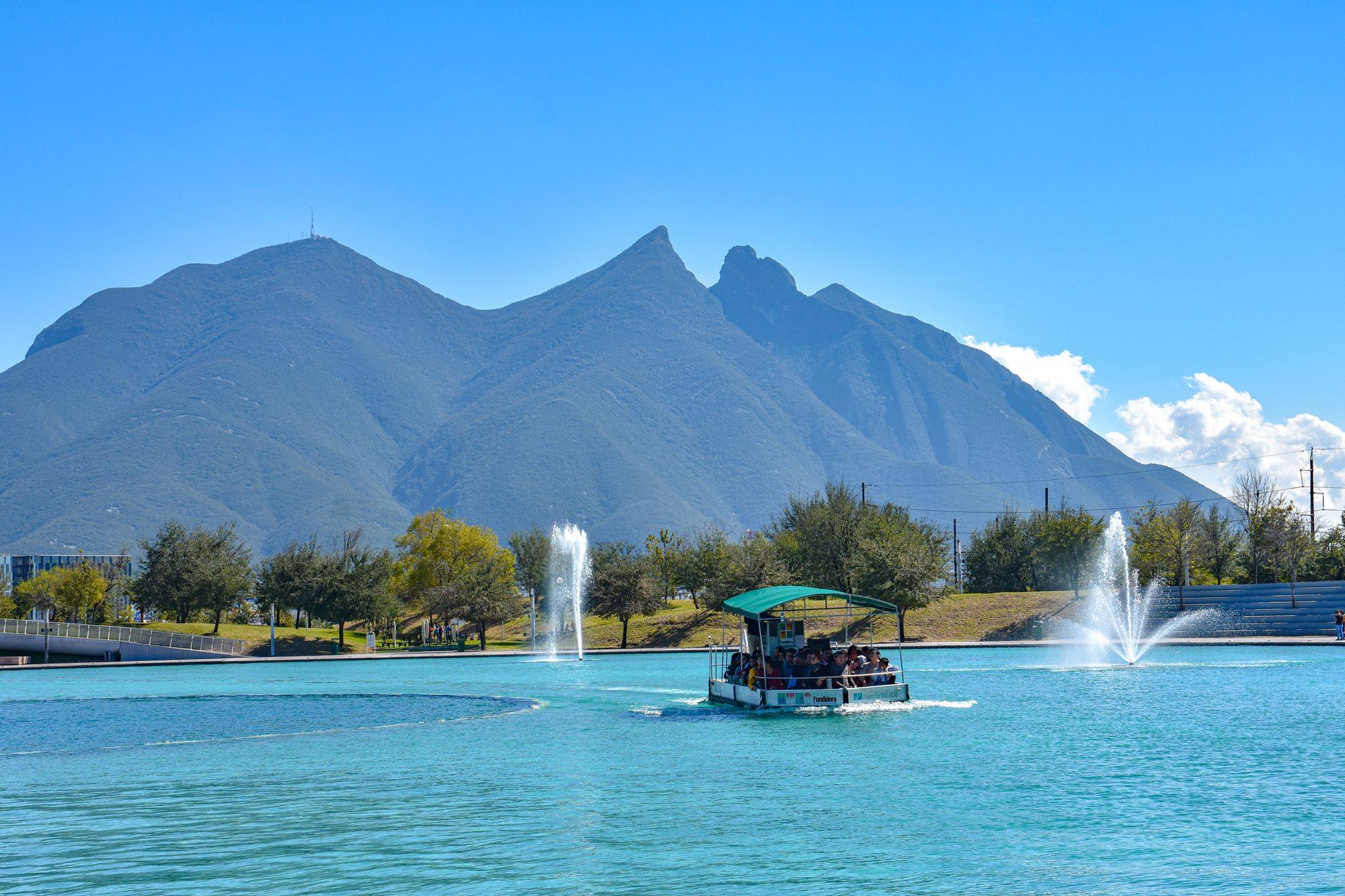 The saddle-shaped Cerro de la Silla above the Paseo Santa Lucia lake in Monterrey, with a boat on the water.