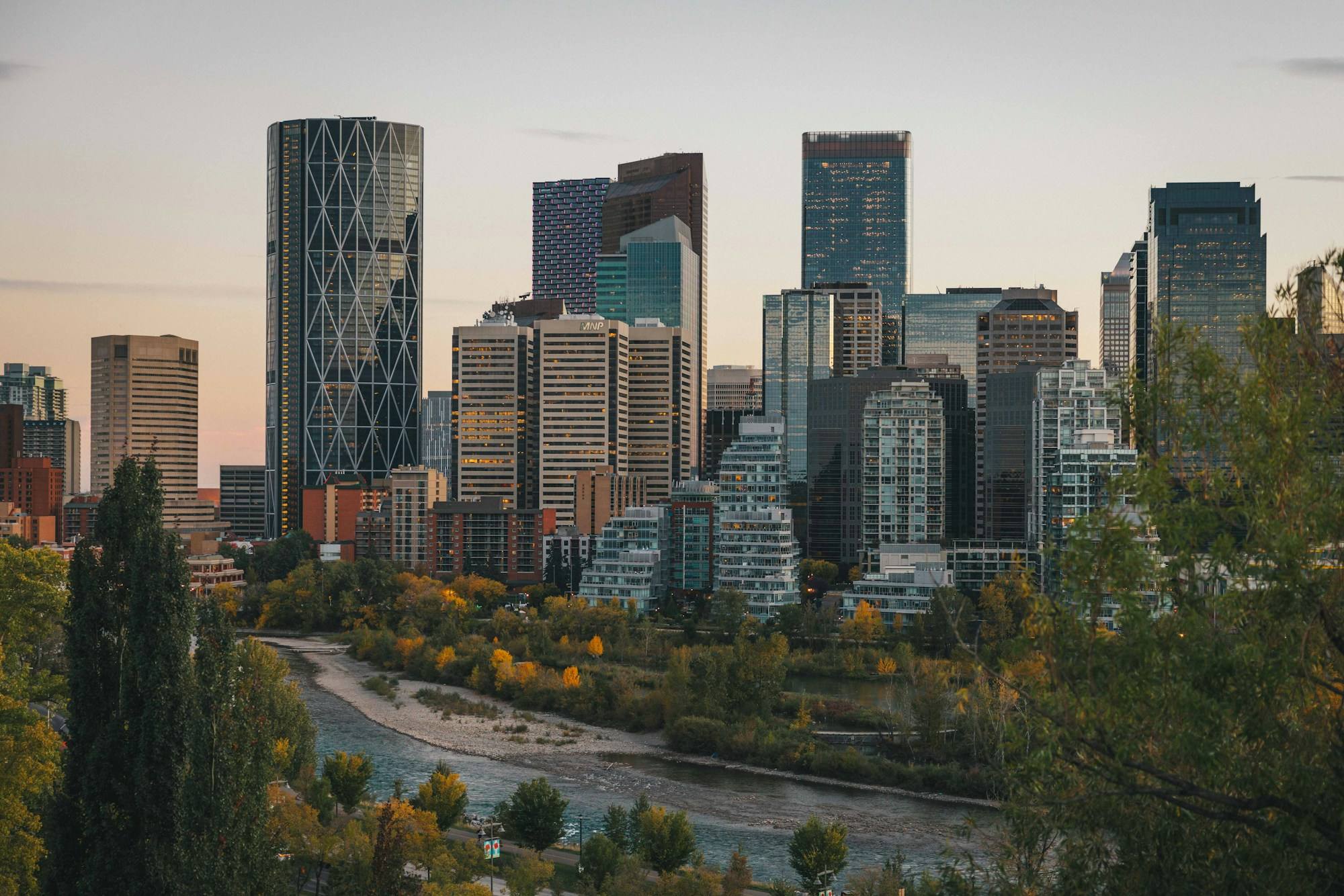 The Calgary skyline at dusk, with The Bow tower and the Bow River winding through autumn trees below it.