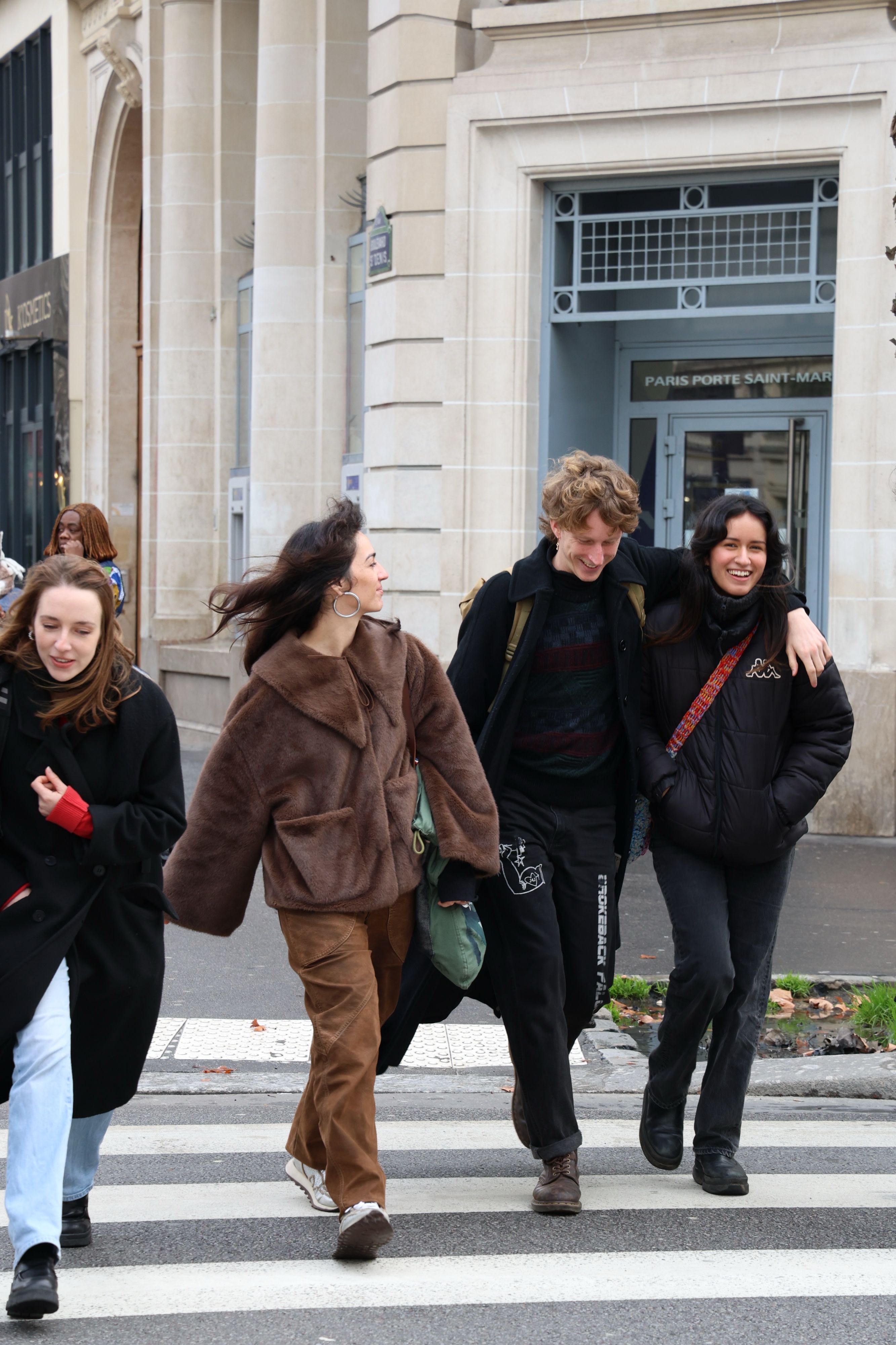 Four people crossing a Paris street together, two of them arm in arm and laughing
