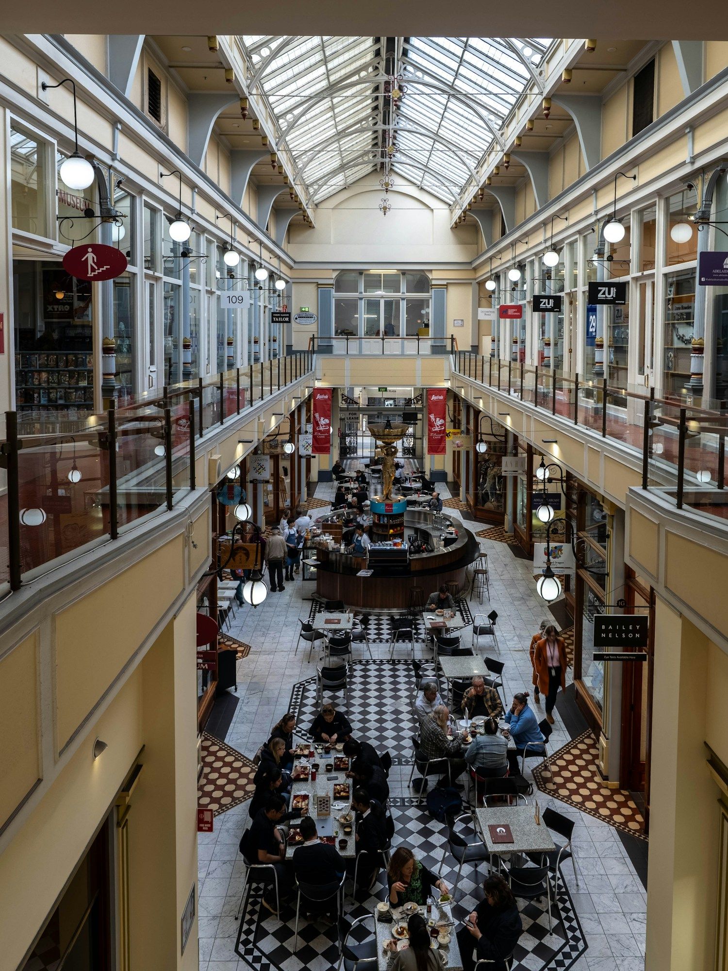 People at café tables inside a historic shopping arcade with a glass roof in Adelaide.