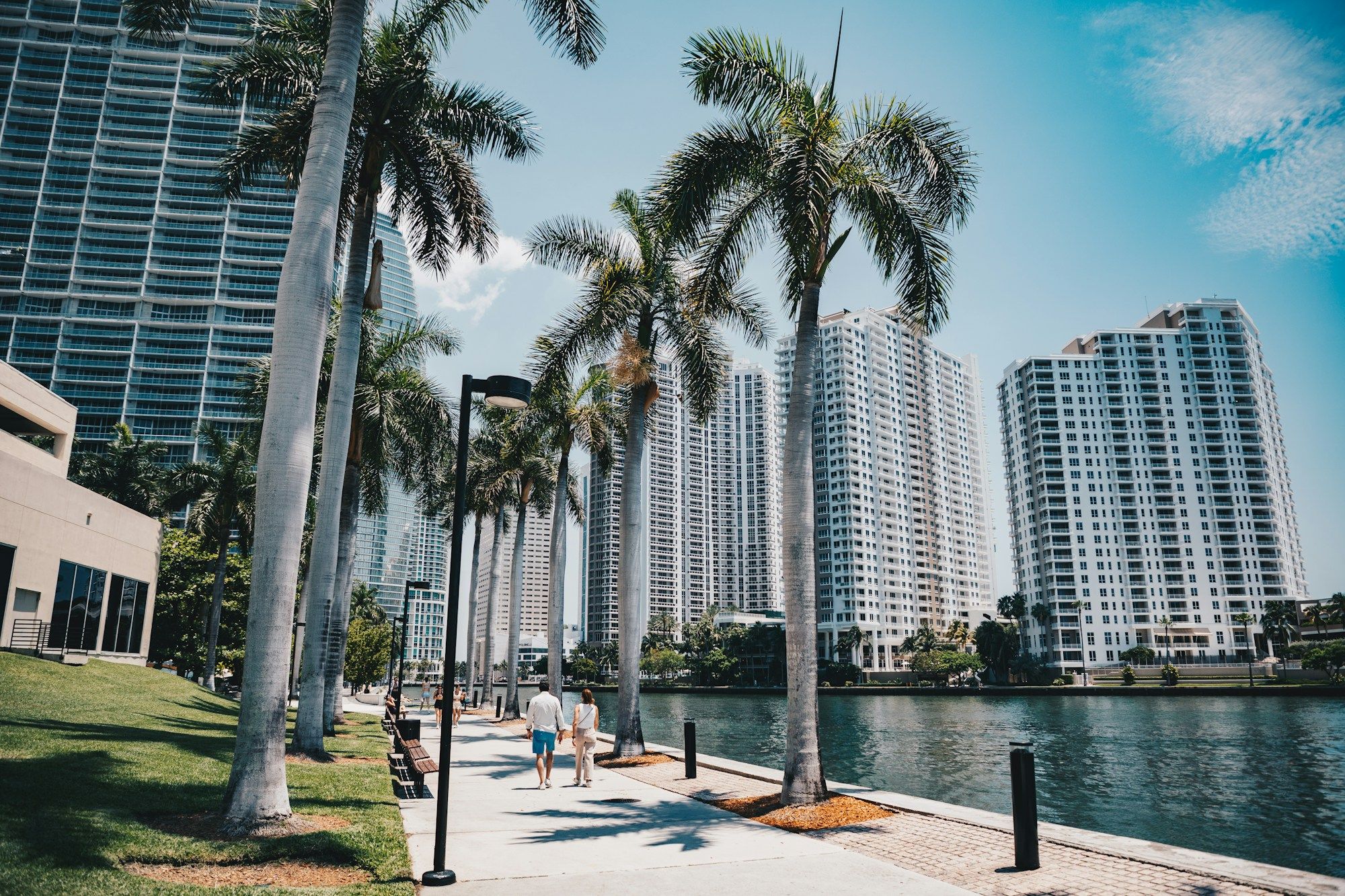 Two people walking along a waterfront path lined with palm trees in Miami, with white high-rise towers across the water.