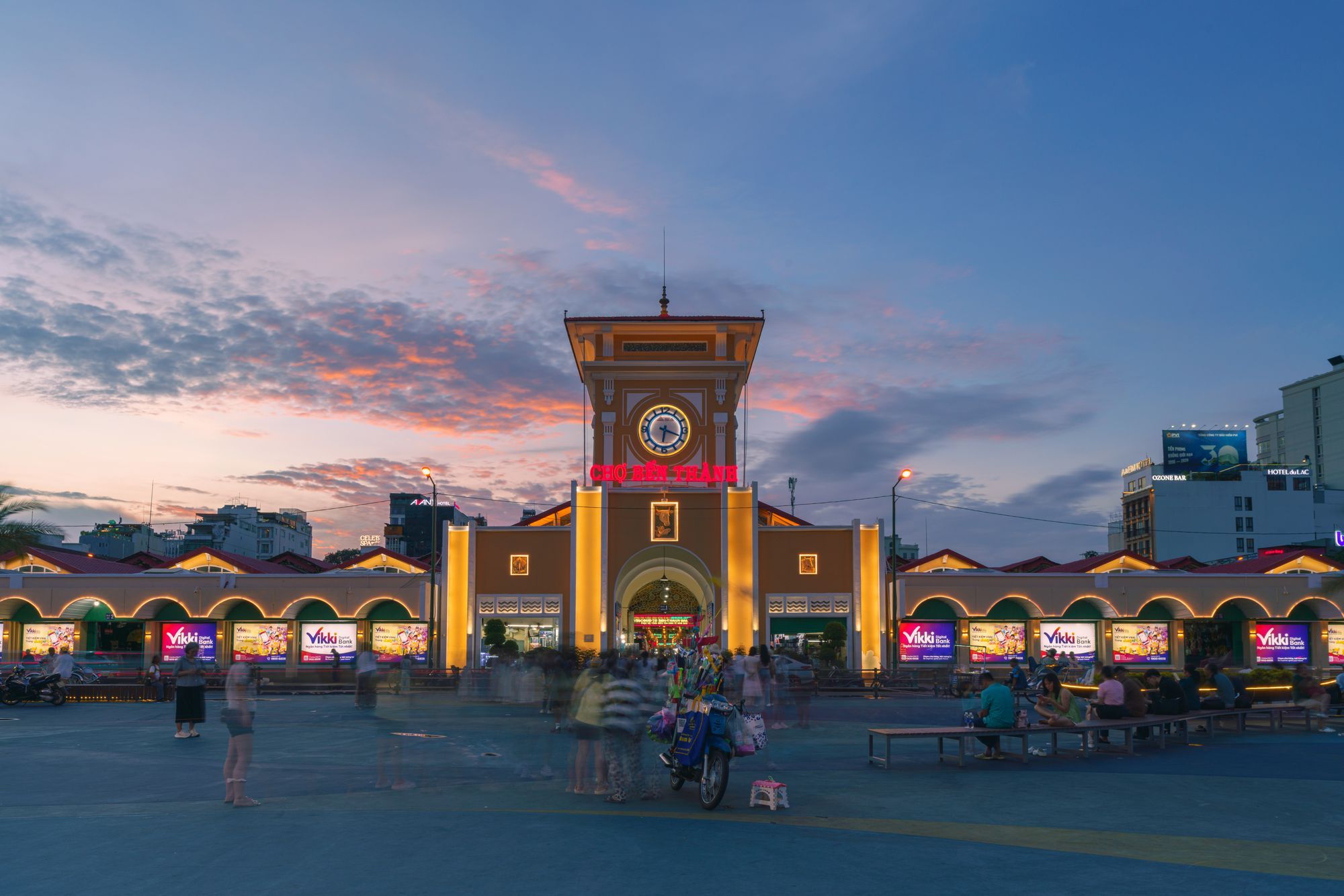 Ben Thanh Market in Ho Chi Minh City at dusk, with its clock tower lit above the entrance and people crossing the square in front.