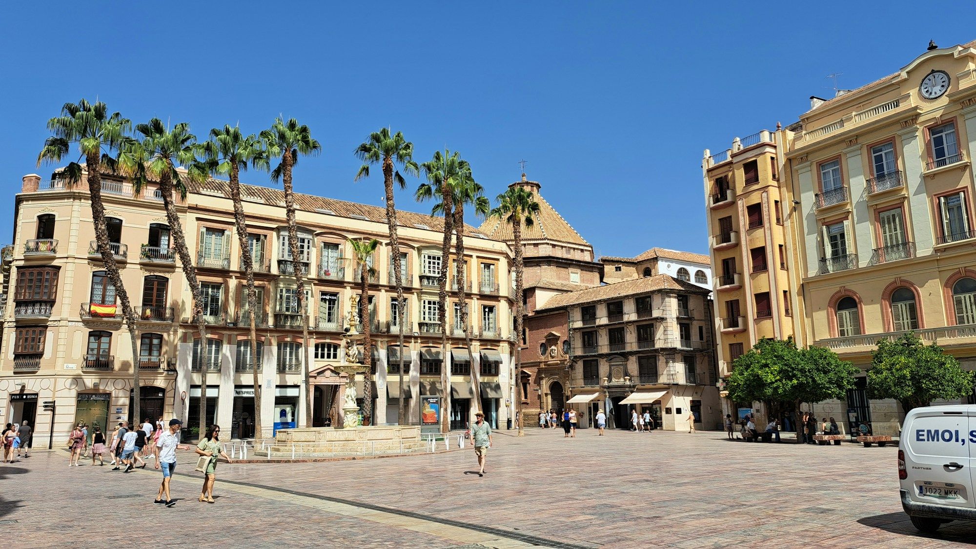 People crossing Plaza de la Constitución in Málaga, with palm trees and old buildings around the square.