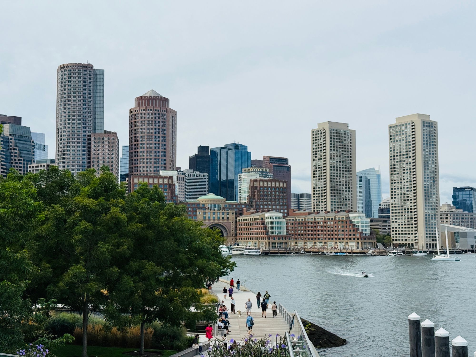 People walking along the Harborwalk beside Boston Harbor, with downtown towers behind the trees.