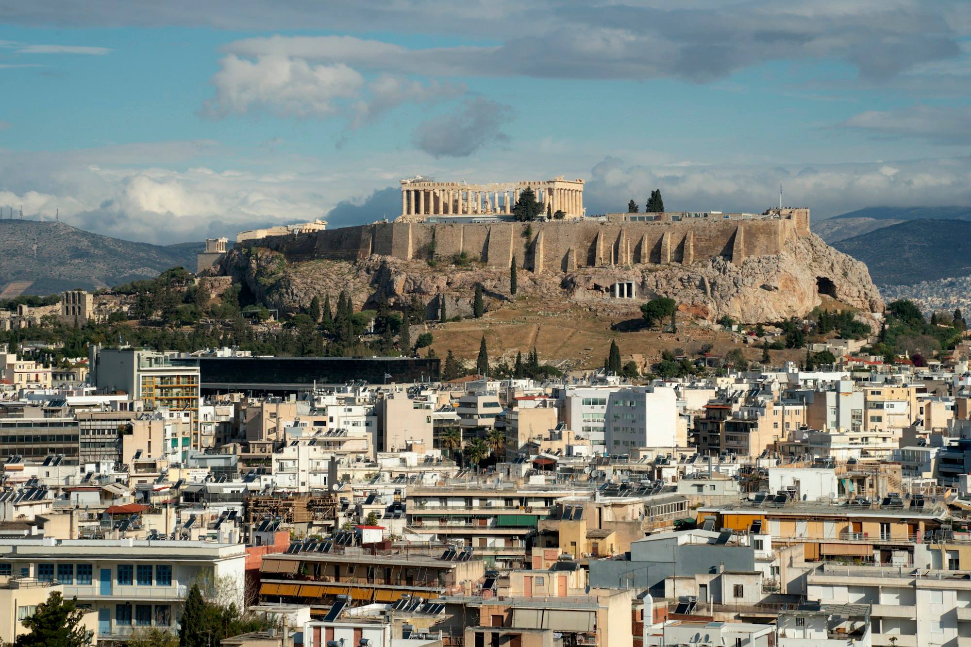 The Acropolis on its rock above the rooftops of central Athens, with hills behind it.