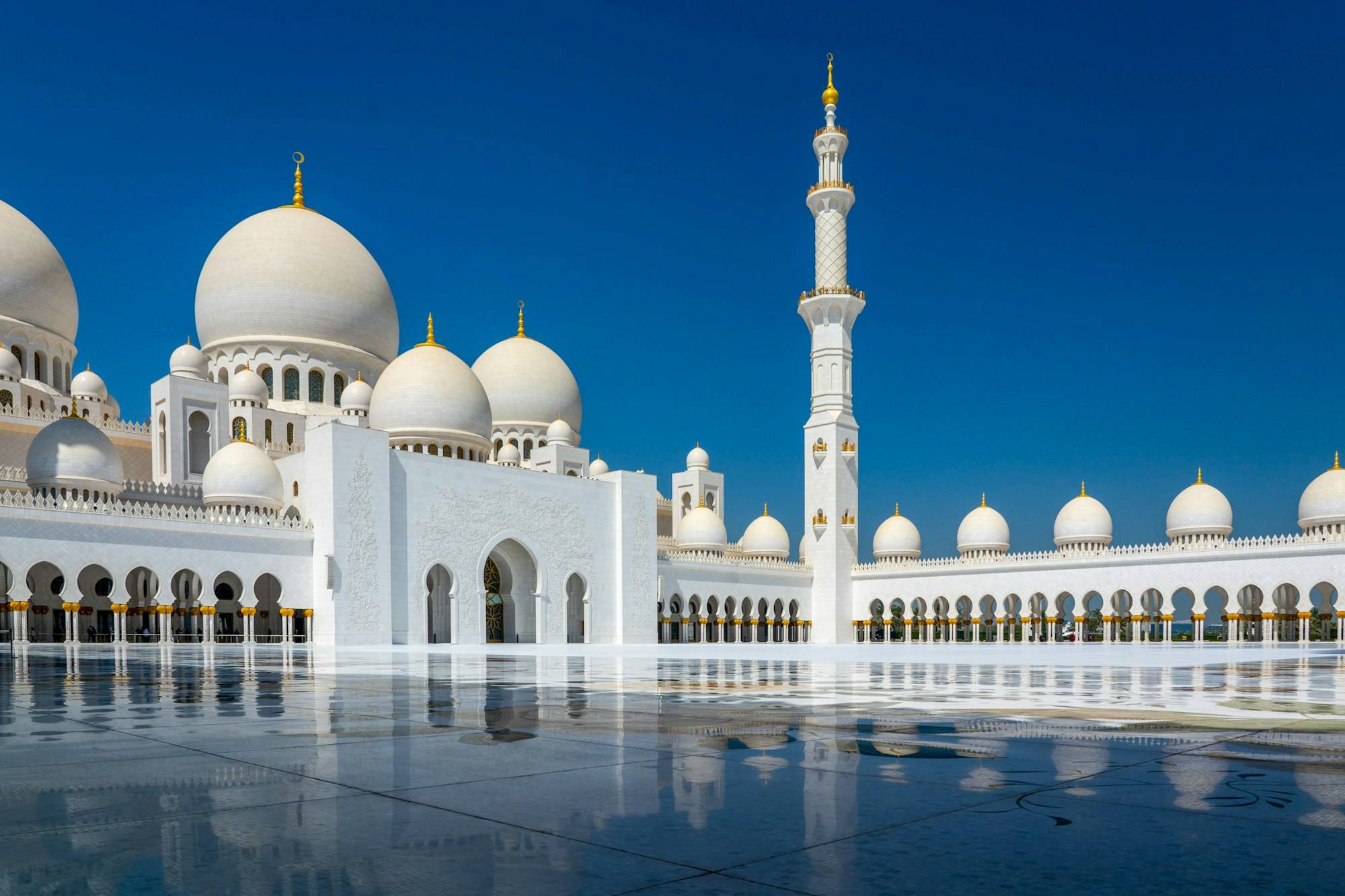 The Sheikh Zayed Grand Mosque in Abu Dhabi, with its white domes and tall minaret against a deep blue sky and reflected in the marble floor.