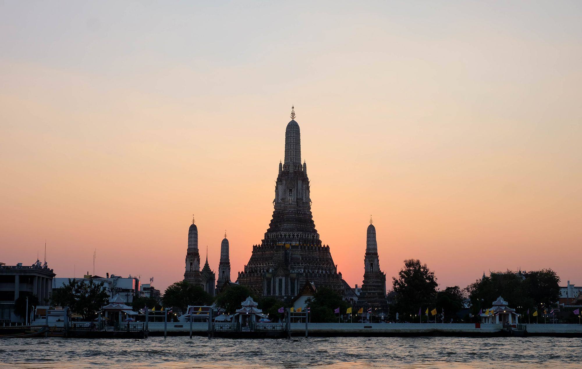 Wat Arun on the bank of the Chao Phraya River in Bangkok at sunset, with the river in the foreground.
