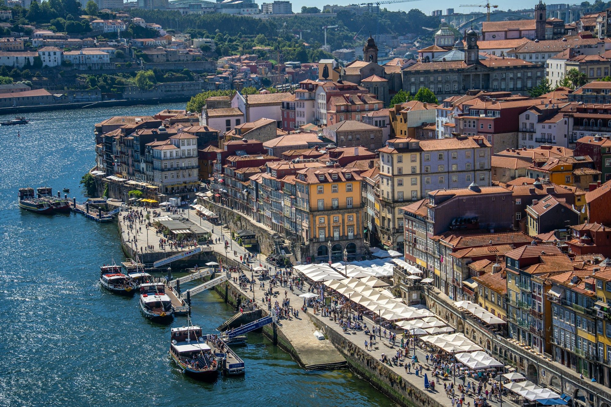 Restaurant terraces and people along the Ribeira riverside in Porto, seen from above, with rabelo boats on the Douro.
