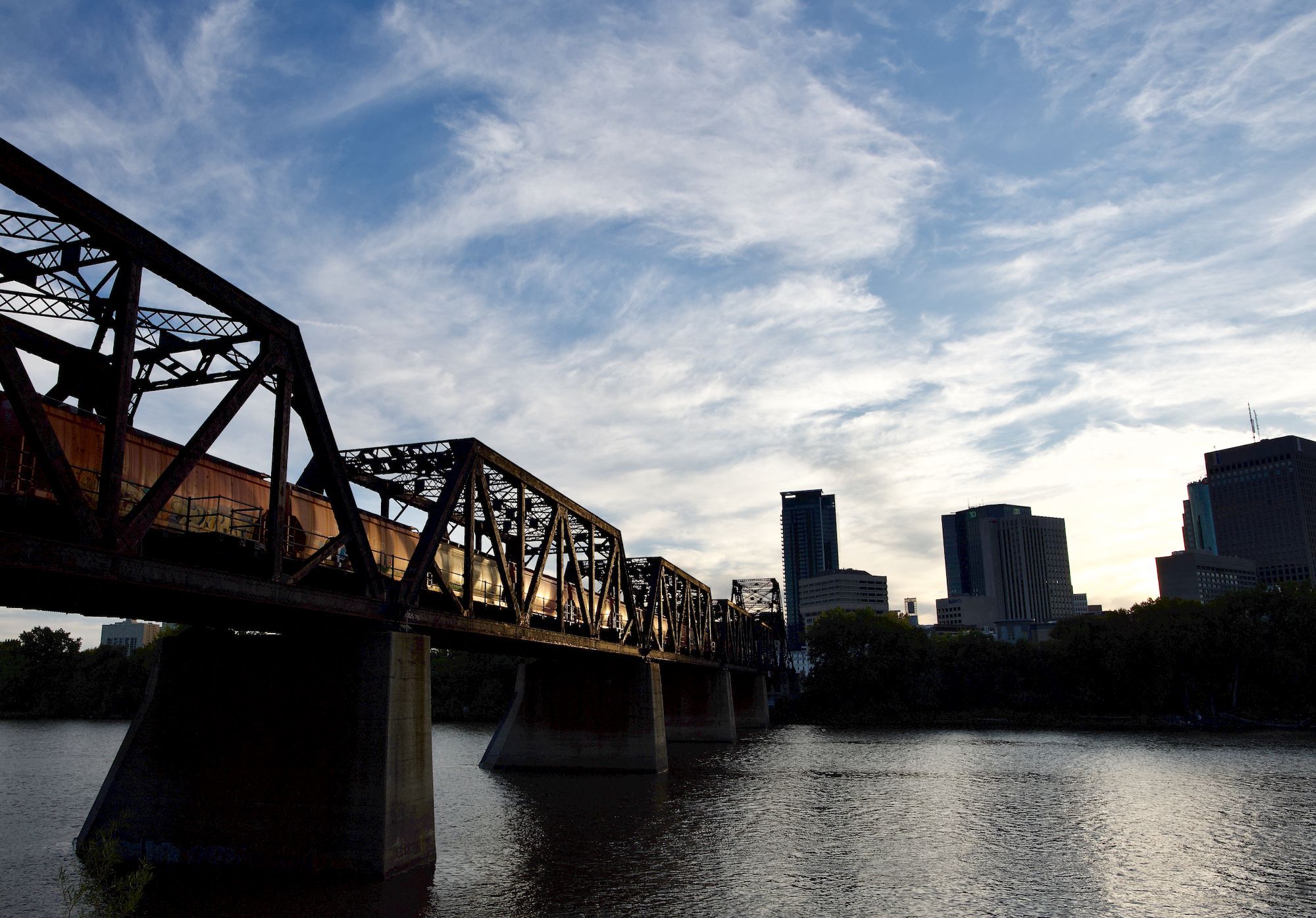 A railway bridge over the river in Winnipeg, with the downtown skyline behind it.