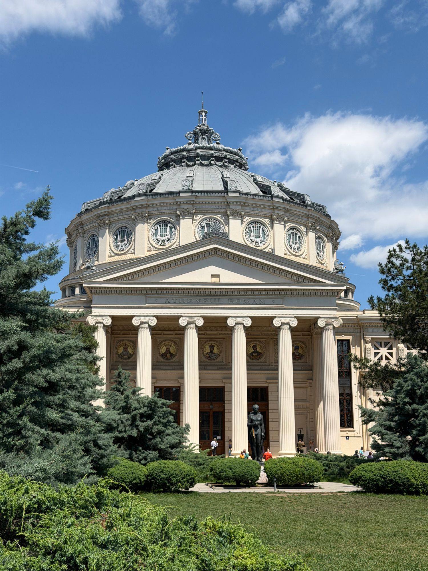 The Romanian Athenaeum in Bucharest, with its dome and columns above a green lawn on a sunny day.