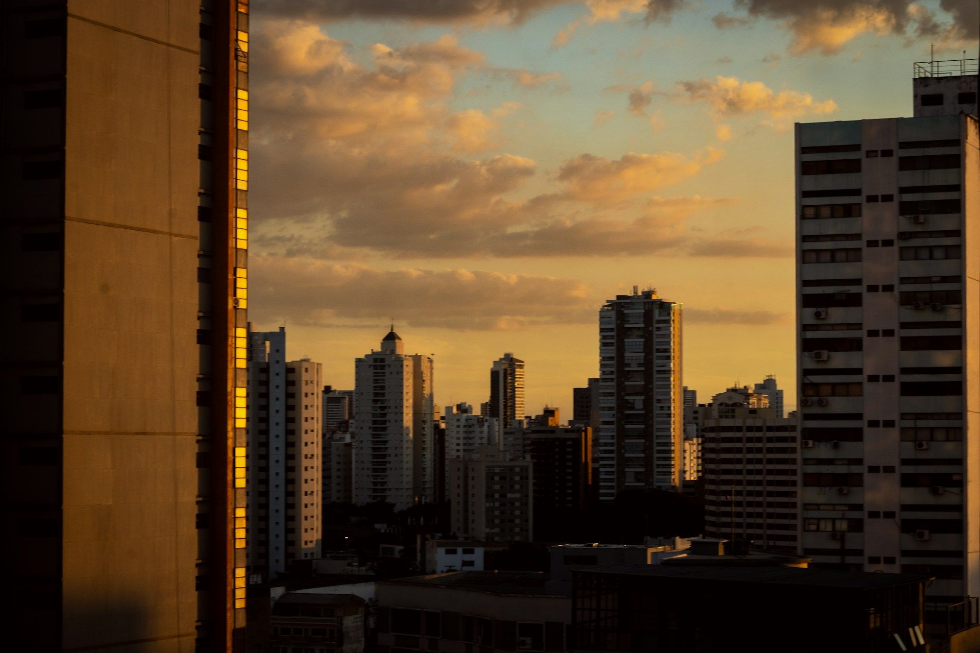 Prédios de Goiânia em silhueta contra nuvens douradas ao pôr do sol, vistos de um andar alto.