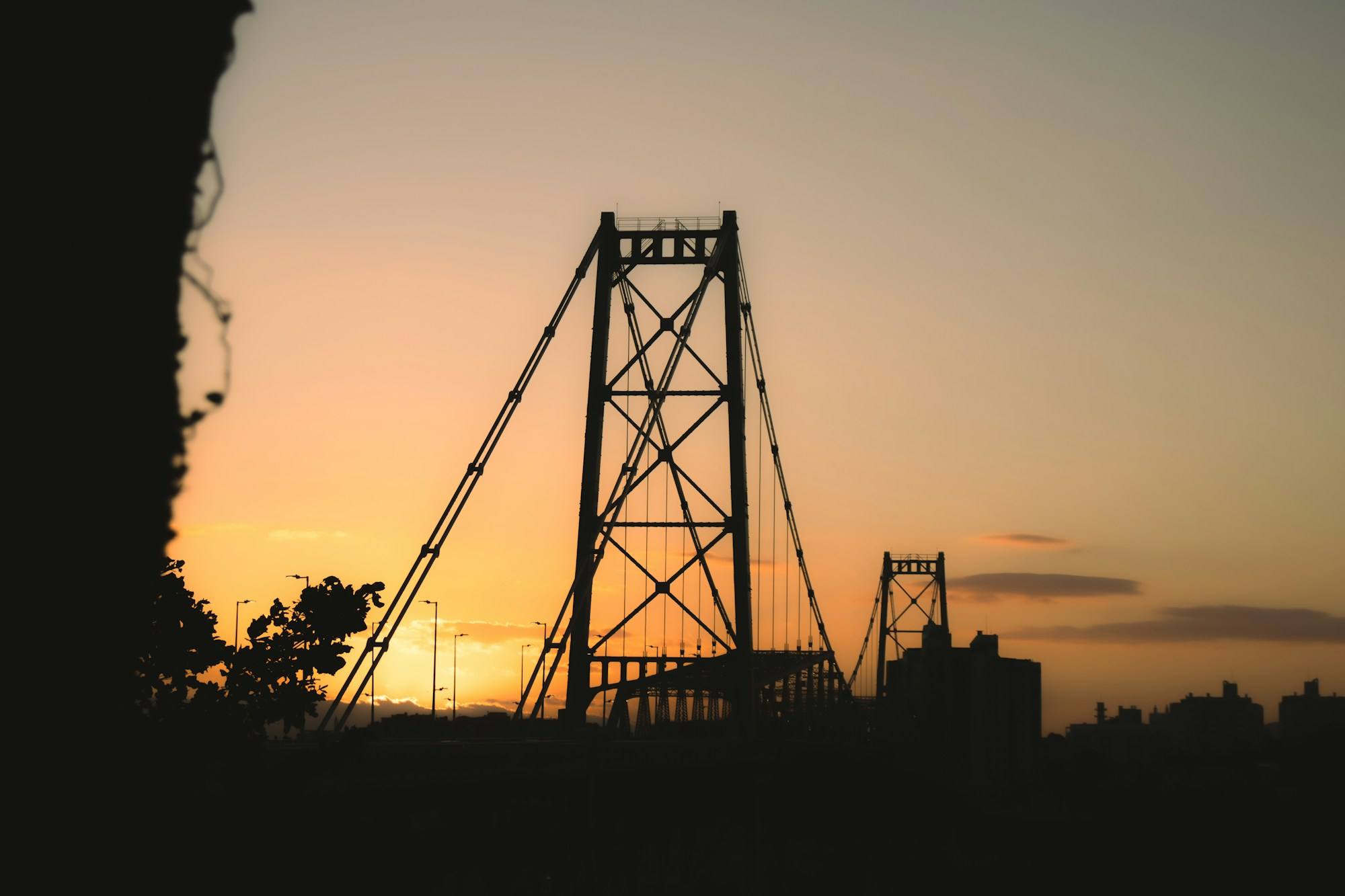 The Hercílio Luz Bridge in Florianópolis in silhouette against an orange sunset sky.