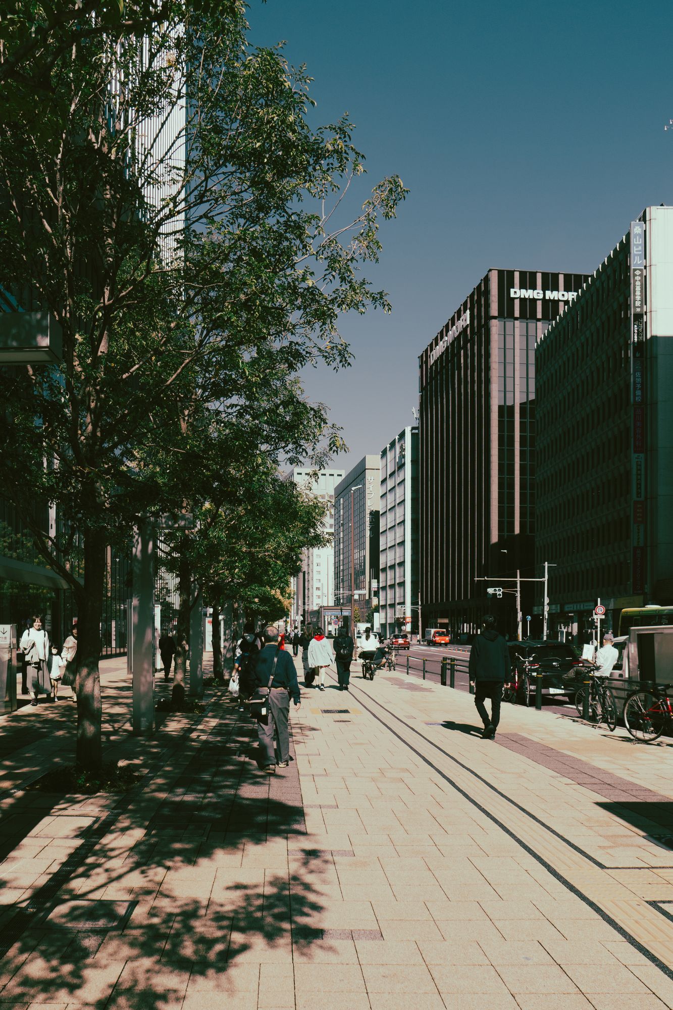 A sunlit sidewalk in Nagoya with people walking between trees and office buildings.
