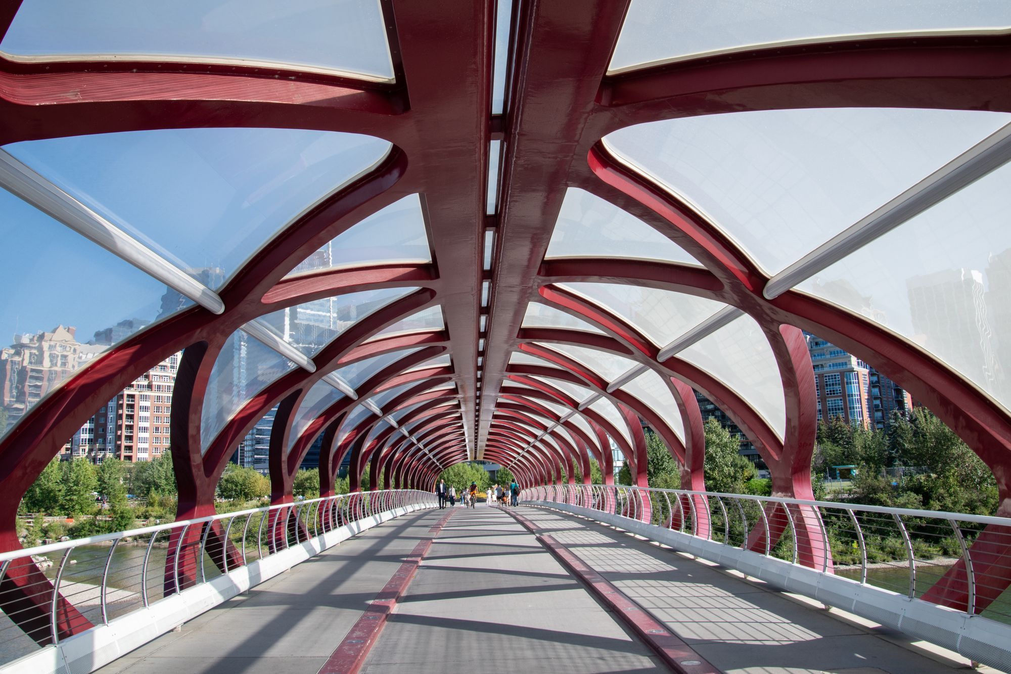 The red steel arches over the walkway of the Peace Bridge in Calgary, with a few people far along it and the Bow River on either side.