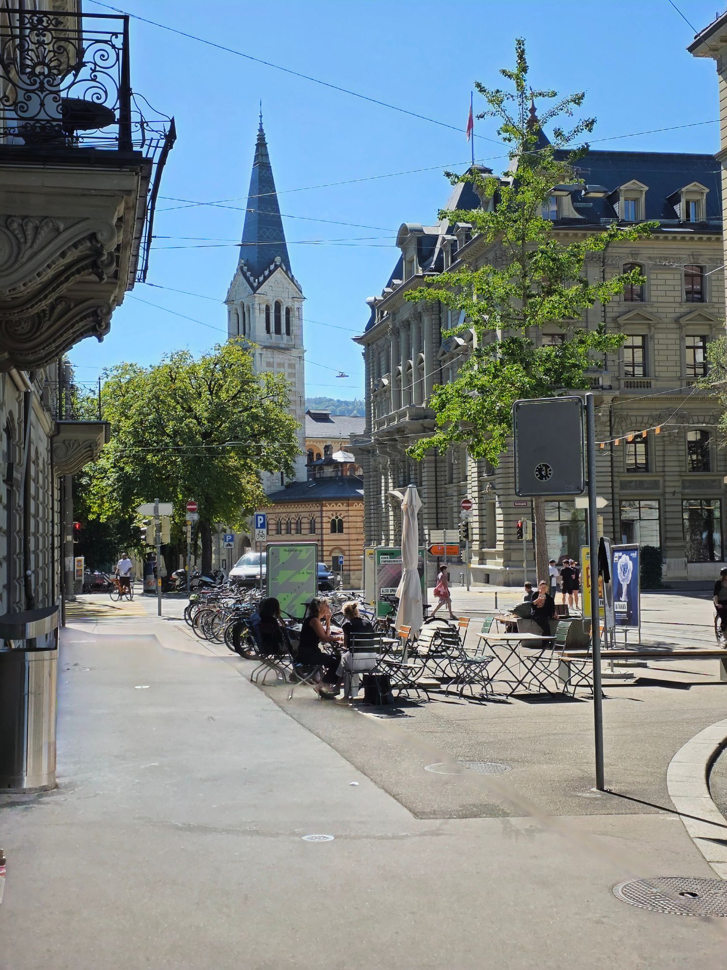 Café terraces with people on a sunny square in Lausanne, with a church spire behind.