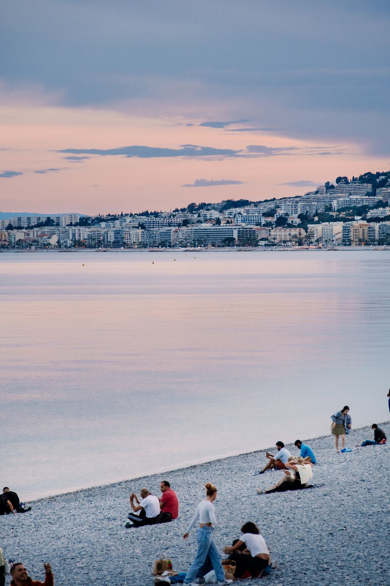 People sitting on the pebble beach in Nice at dusk, with the city along the shore in the distance.