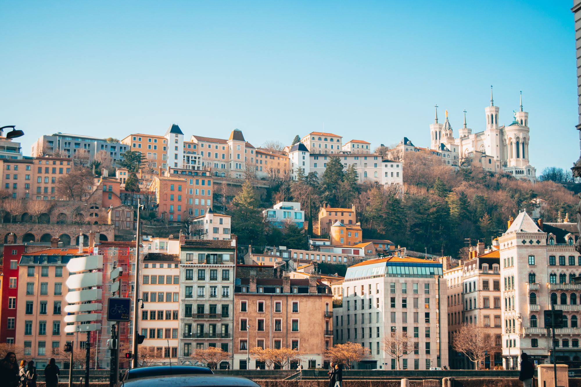 The Basilica of Notre-Dame de Fourvière on its wooded hill above the buildings of Lyon, under a blue sky.