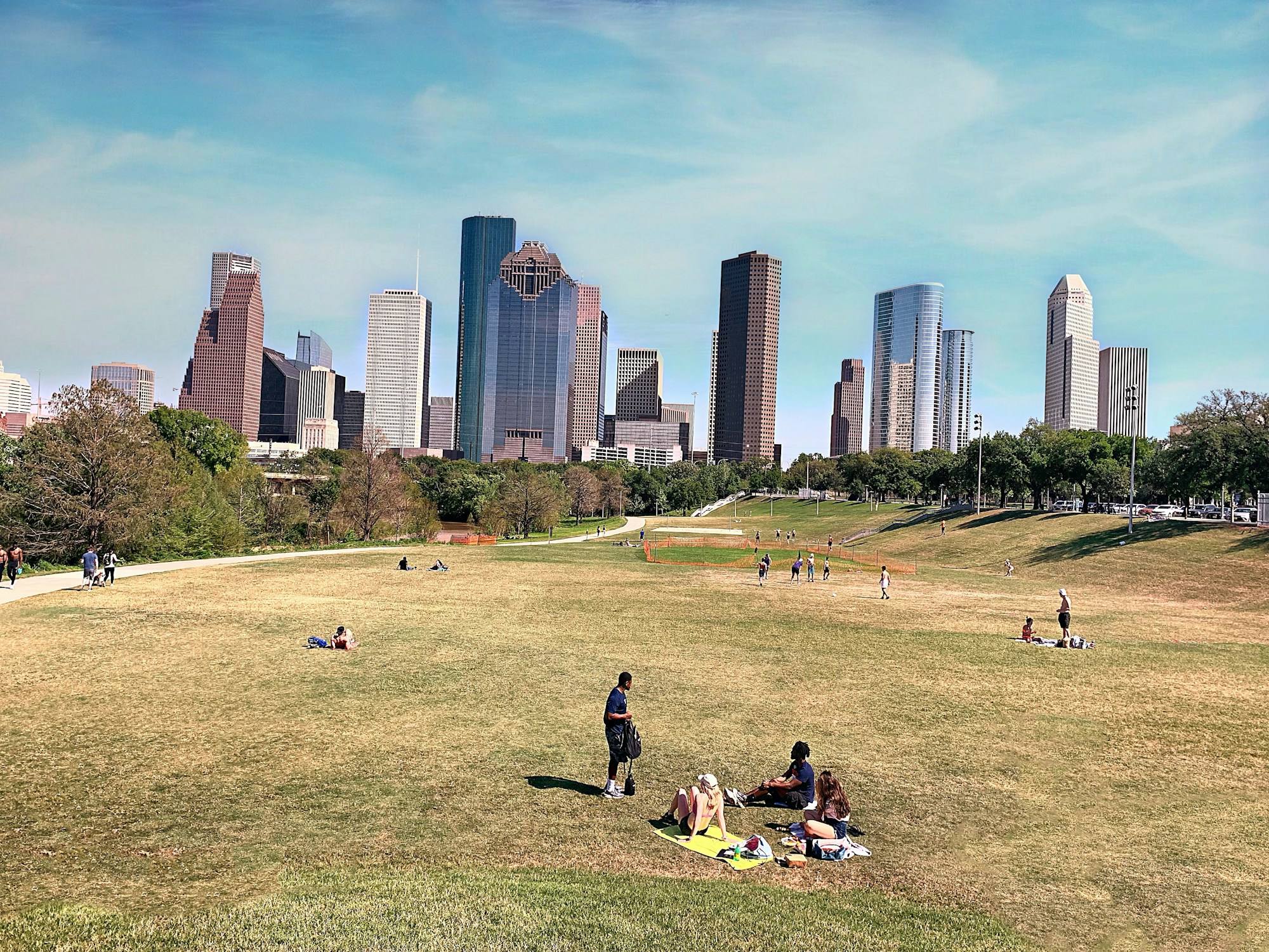 The downtown Houston skyline above the lawns of Buffalo Bayou Park, with a few people sitting on the grass on a sunny day