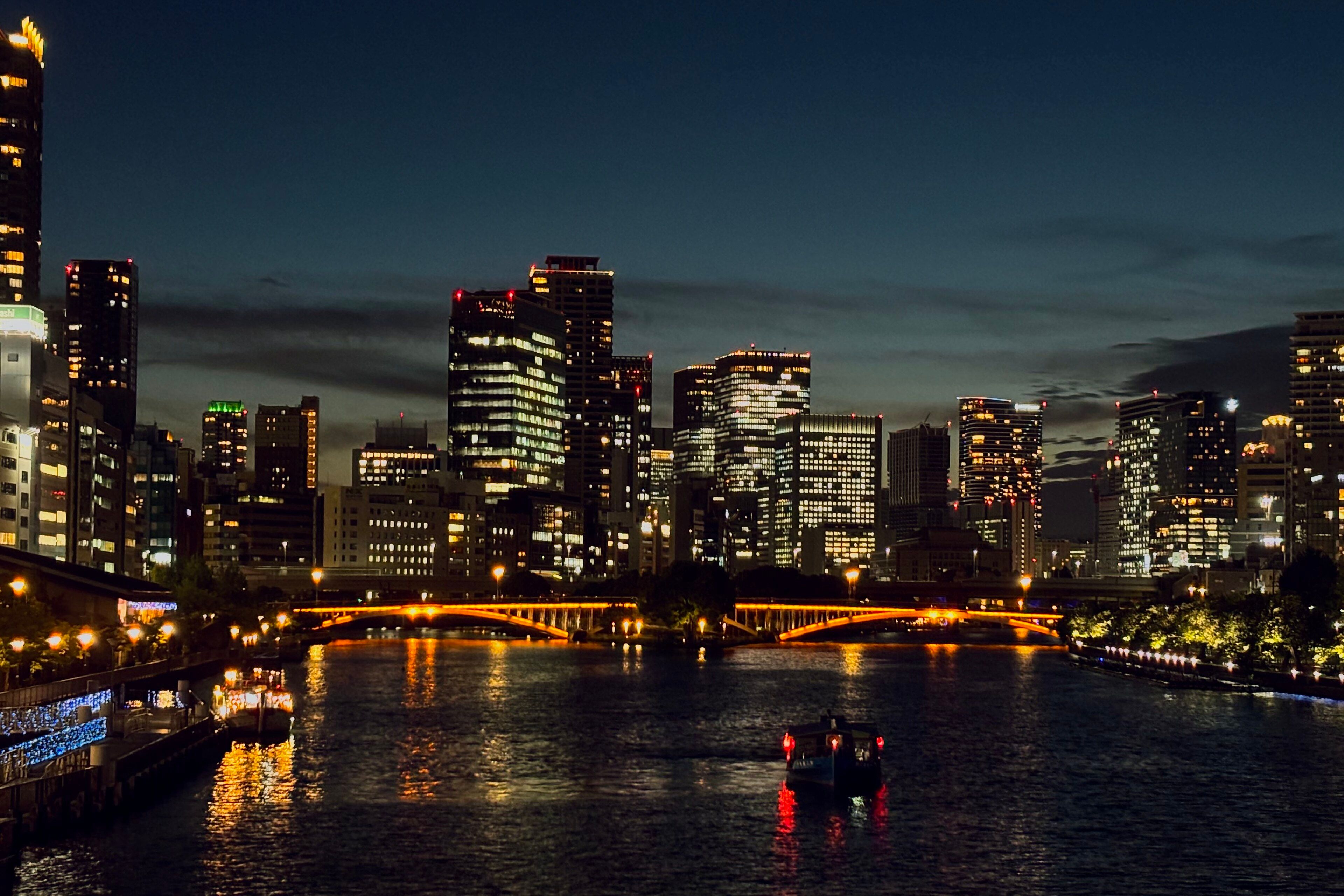 Towers and bridges along the Nakanoshima riverfront in Osaka lit up at night, with a boat on the river.