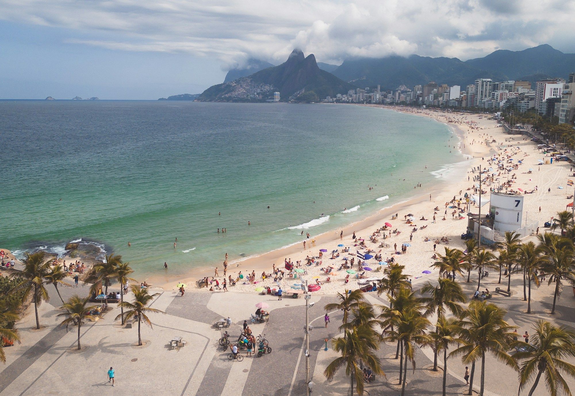 Gente caminhando e pedalando no calçadão de Ipanema, no Rio de Janeiro, com a praia e o Morro Dois Irmãos ao fundo.