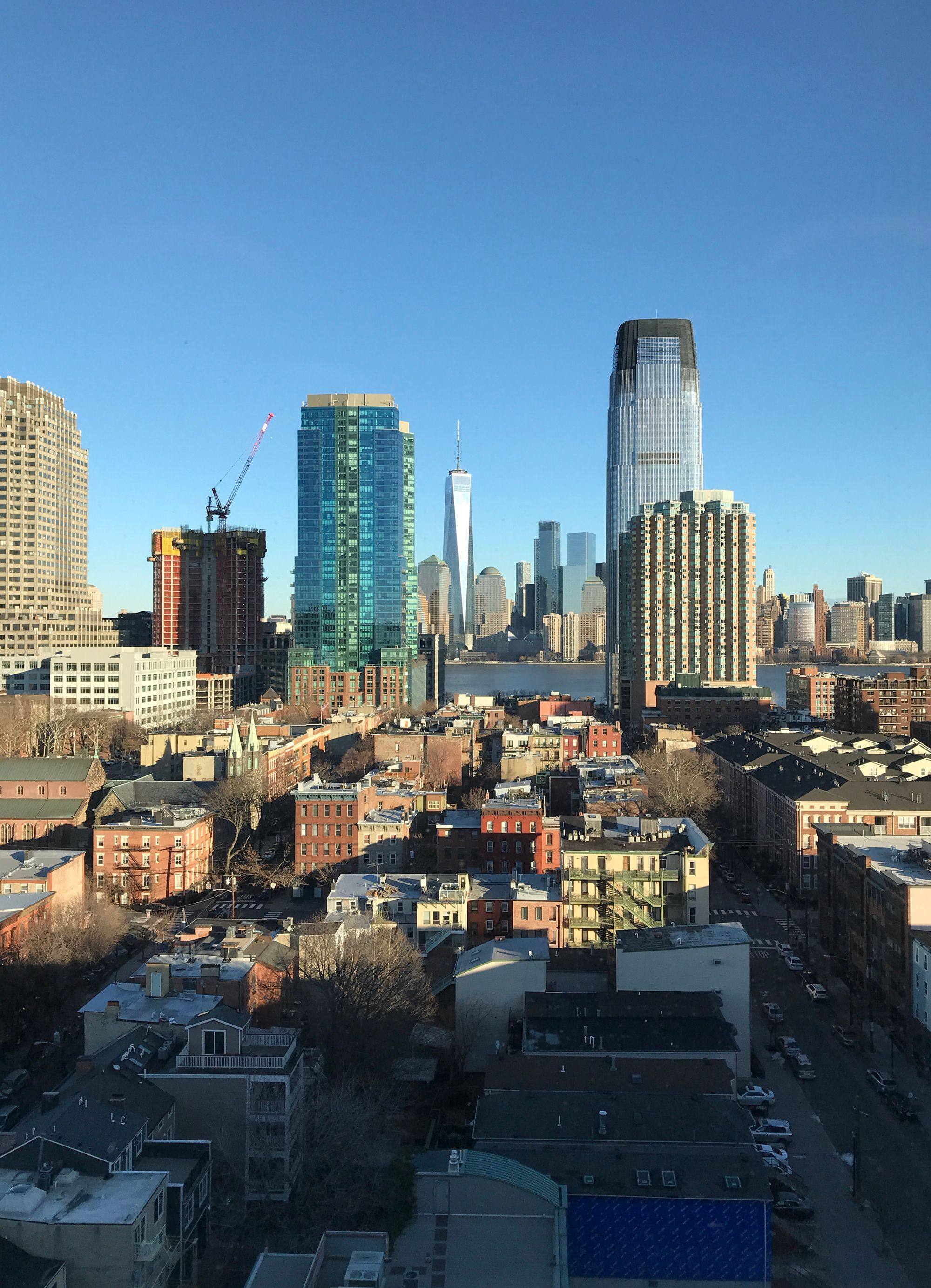 Skyscrapers in downtown Jersey City, with One World Trade Center visible behind across the Hudson.