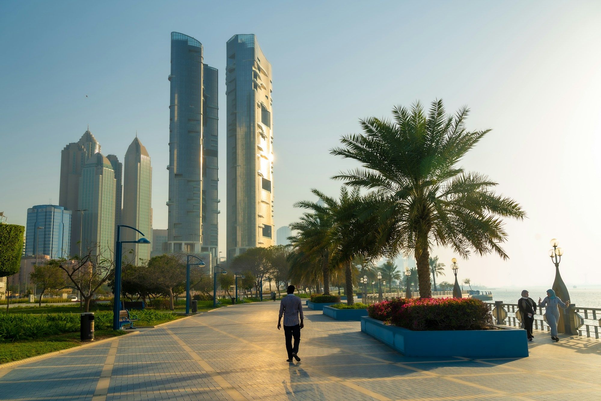 People walking along the Corniche promenade in Abu Dhabi at sunset, with palm trees, flower beds and tall towers behind.