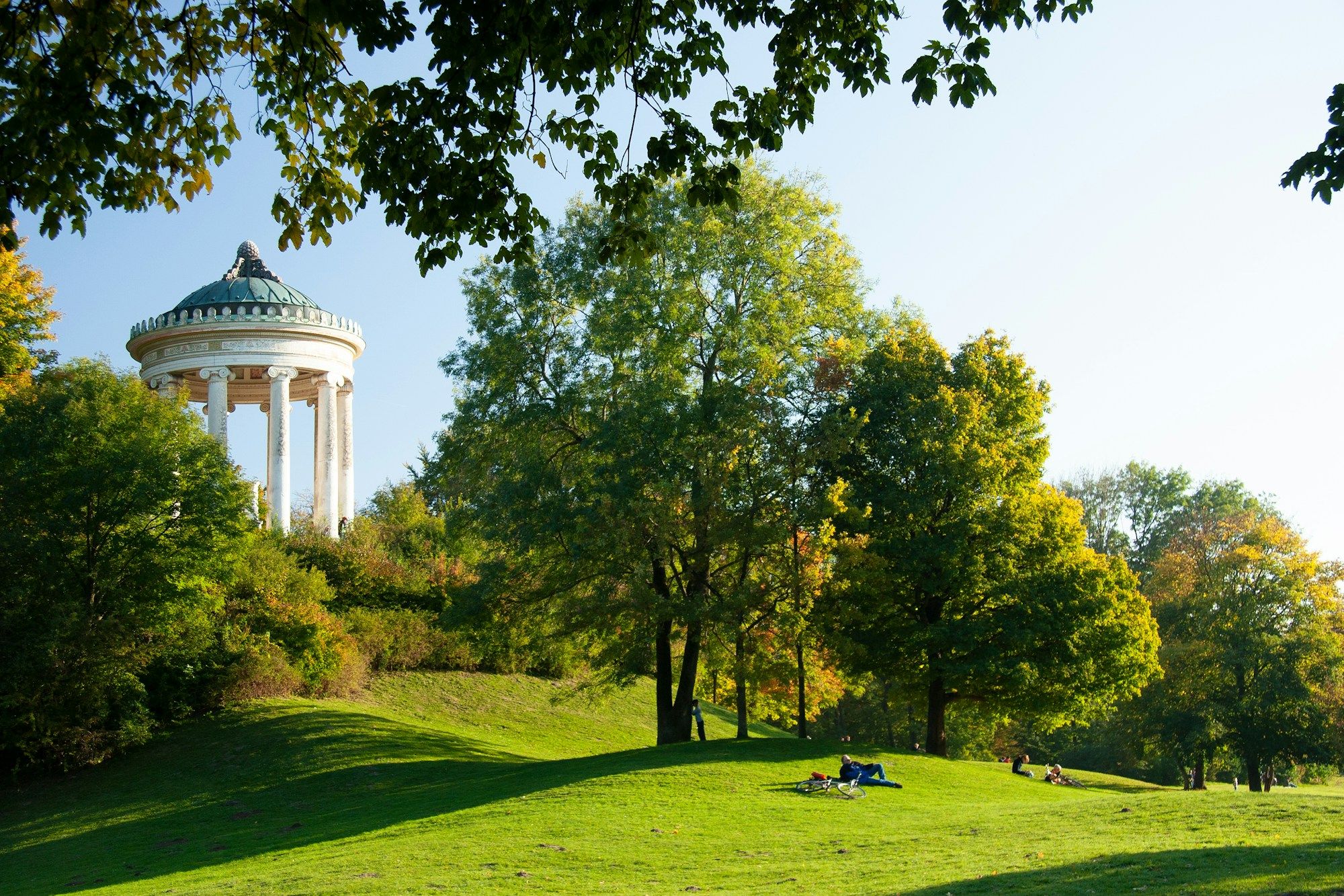 The Monopteros temple above a sunny meadow in the Englischer Garten in Munich, with a few people resting on the grass.
