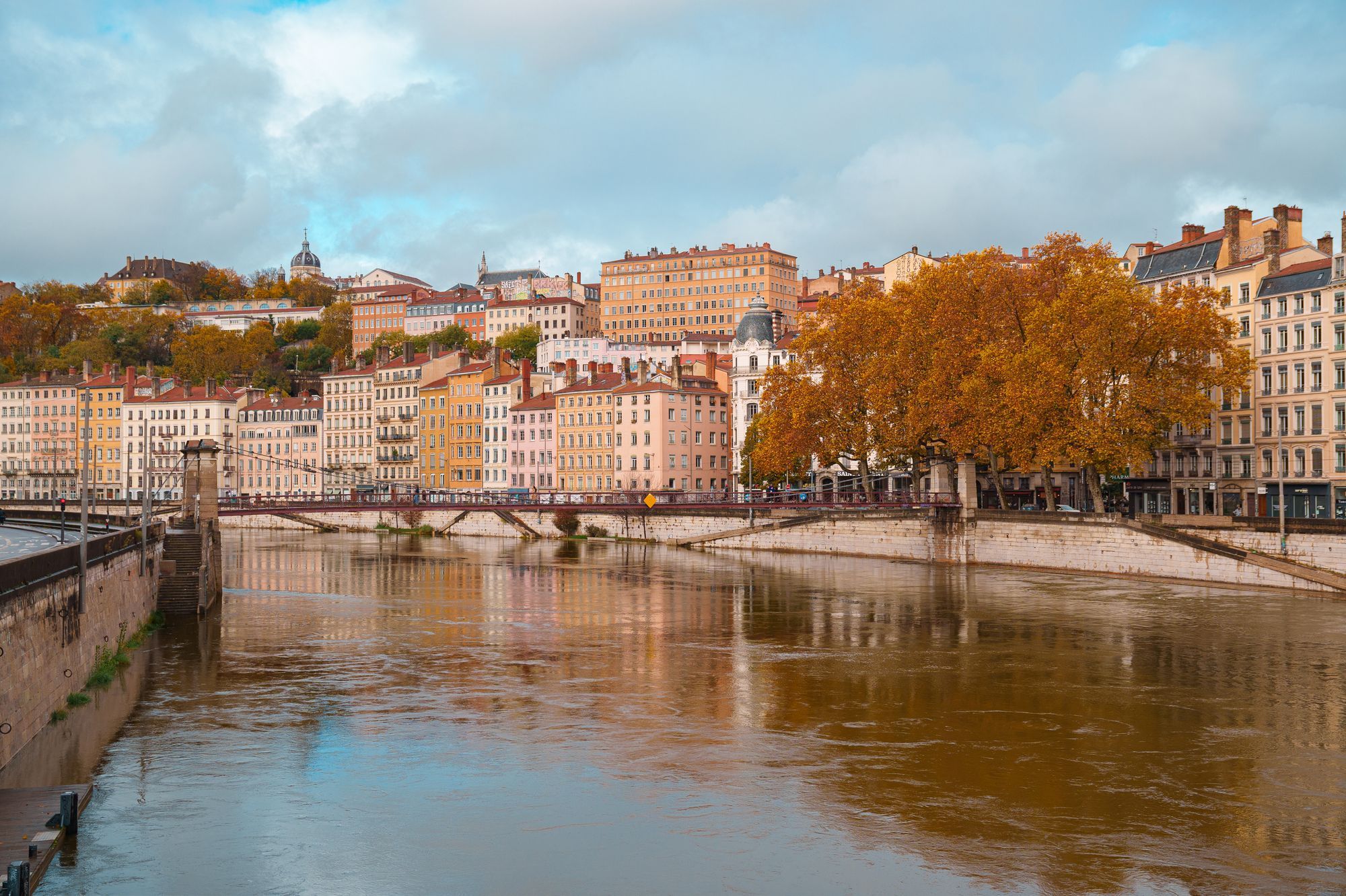 The Saône river in Lyon in autumn, with an orange tree and the pastel quayside buildings of Vieux Lyon across the water.