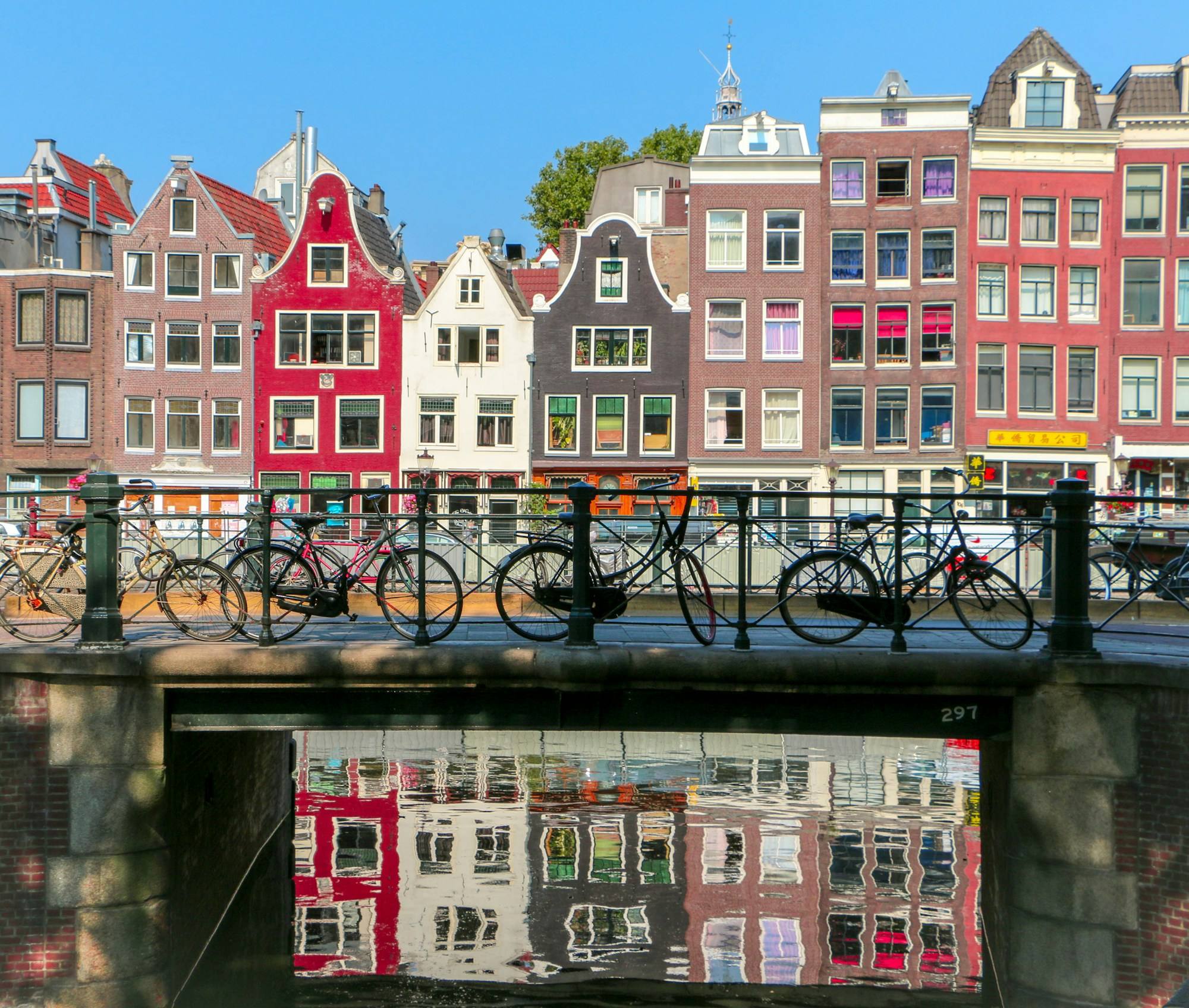 Gabled canal houses behind a bridge railing lined with bicycles in Amsterdam, reflected in the canal below.