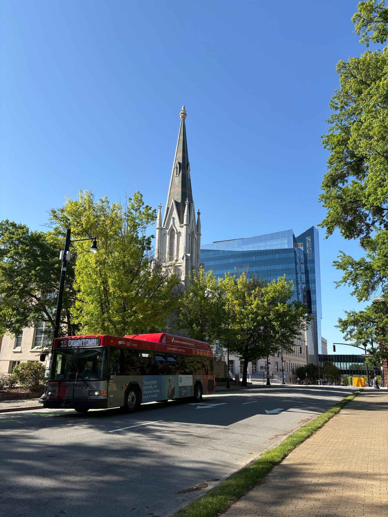 A downtown Raleigh street with a bus, a church spire and a glass office tower behind trees.