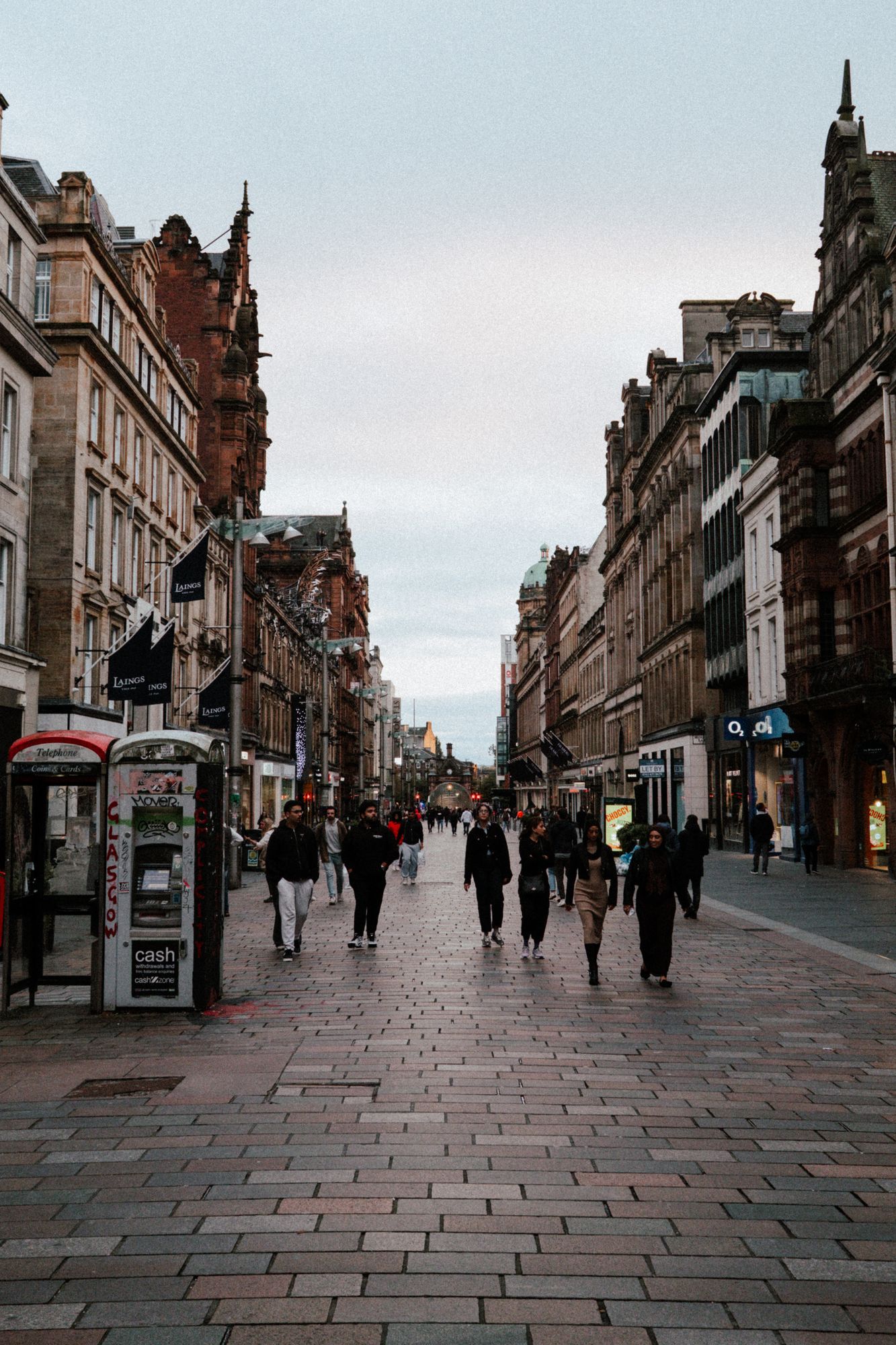 People walking down a pedestrian street lined with sandstone buildings in central Glasgow at dusk.